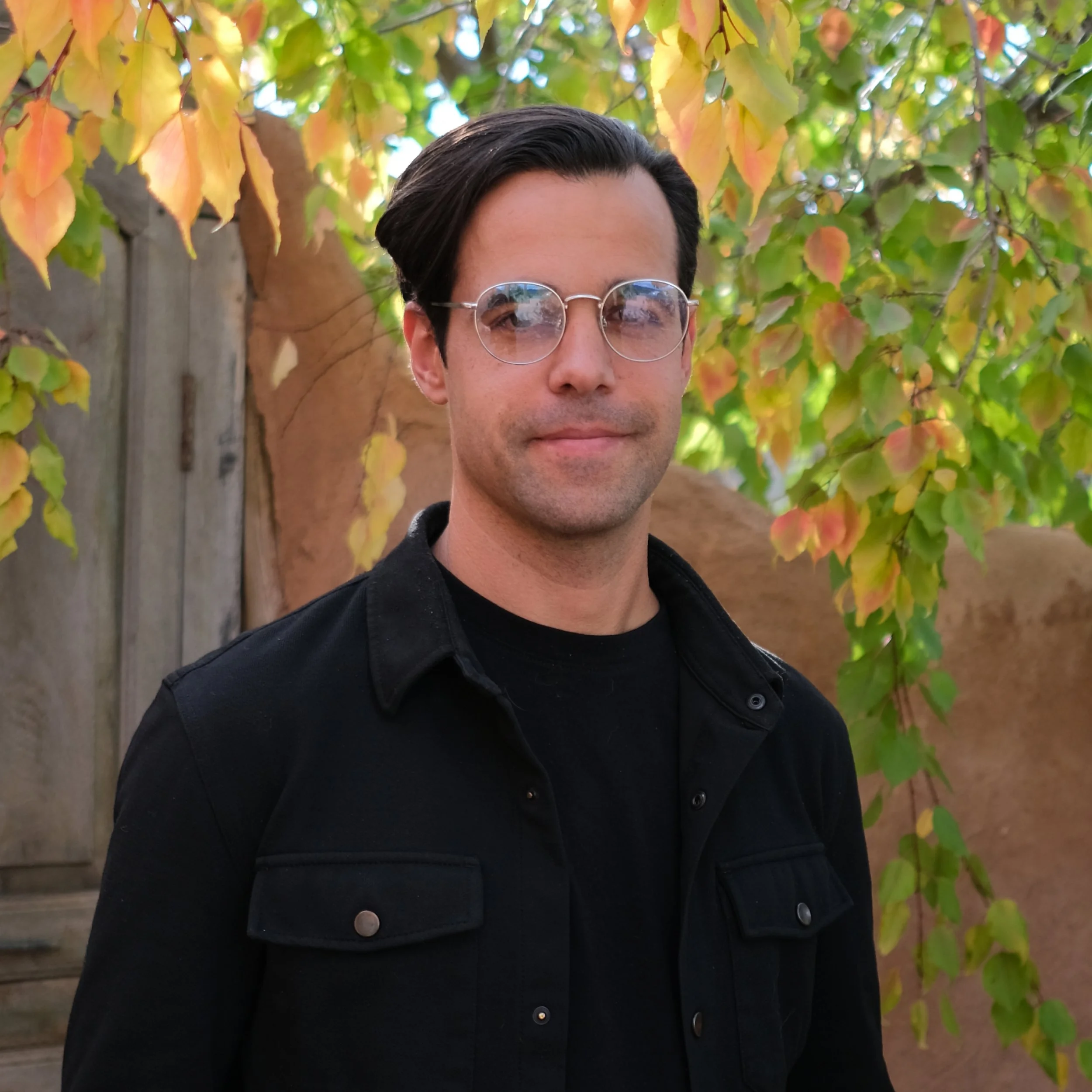 A man with dark hair and glasses standing outdoors in front of a tree with colorful autumn leaves.