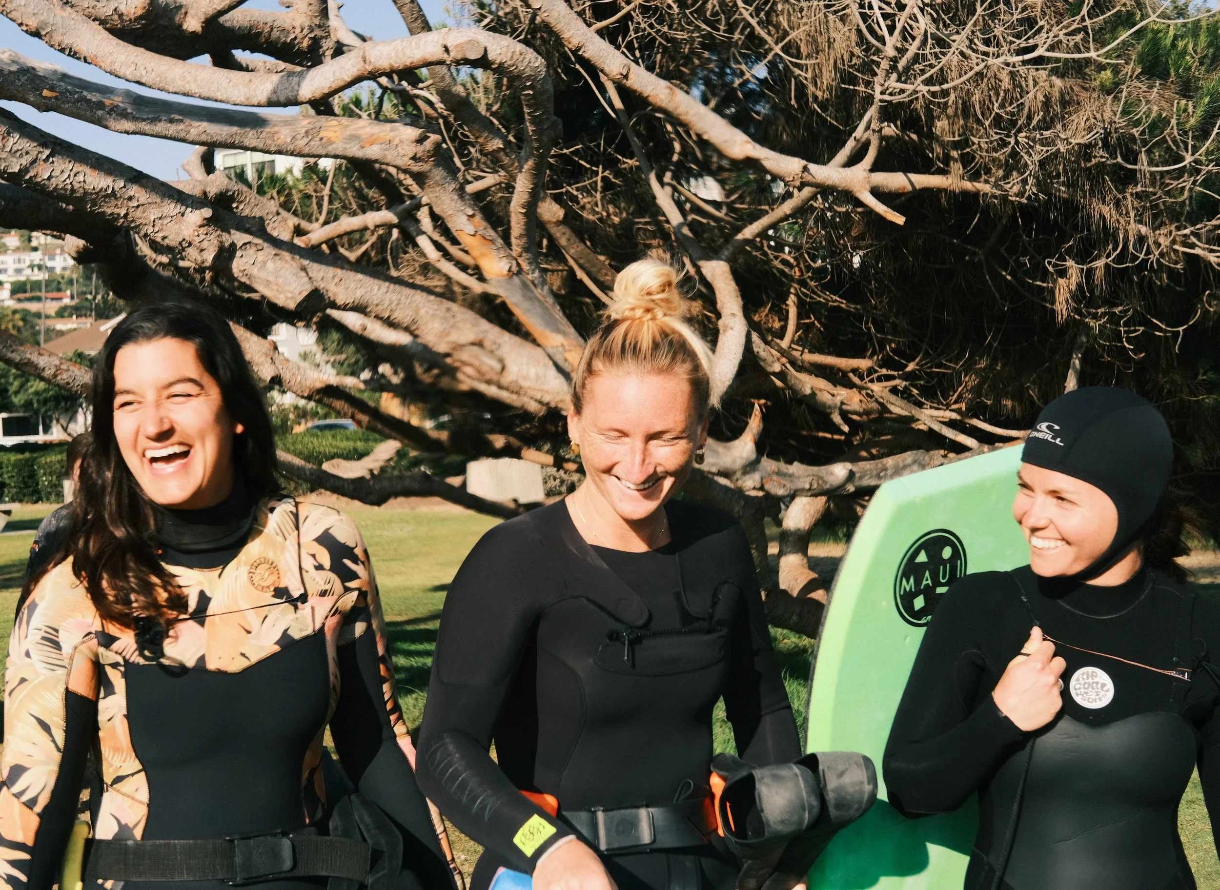Three women in wetsuits standing outdoors near a large tree, smiling and laughing.