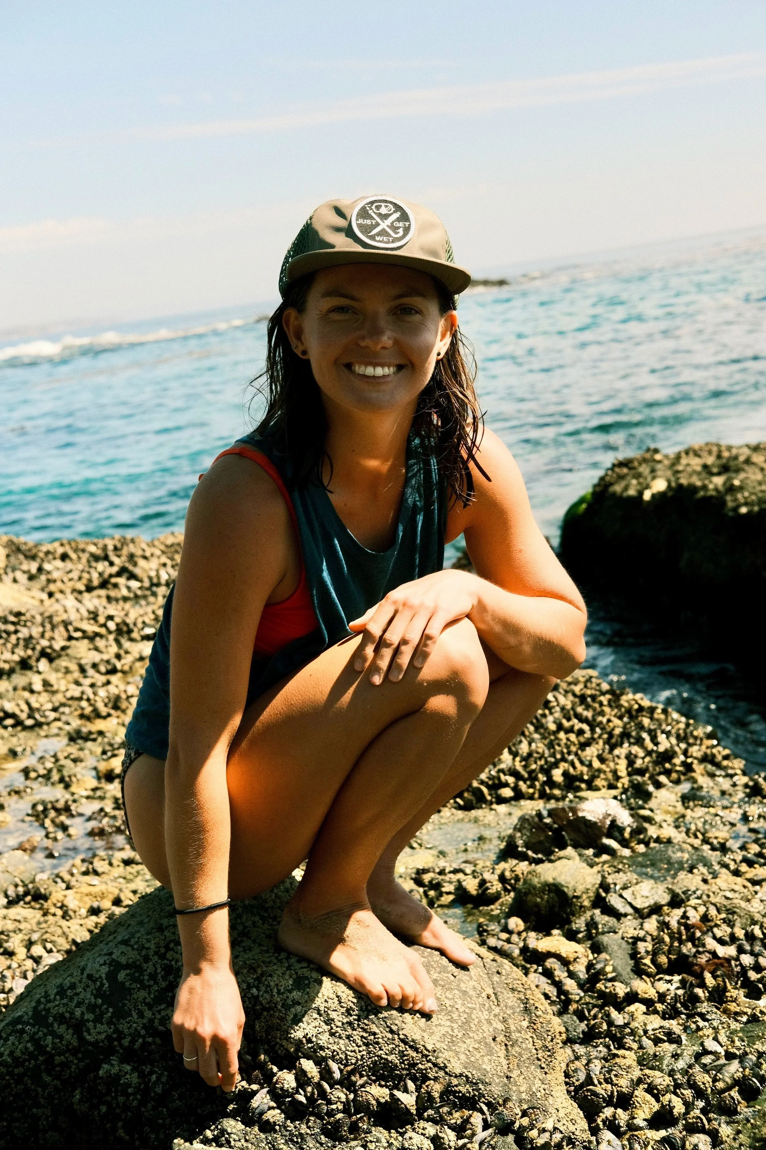 A woman squatting on a rocky shoreline, smiling at the camera, with the ocean and sky in the background.