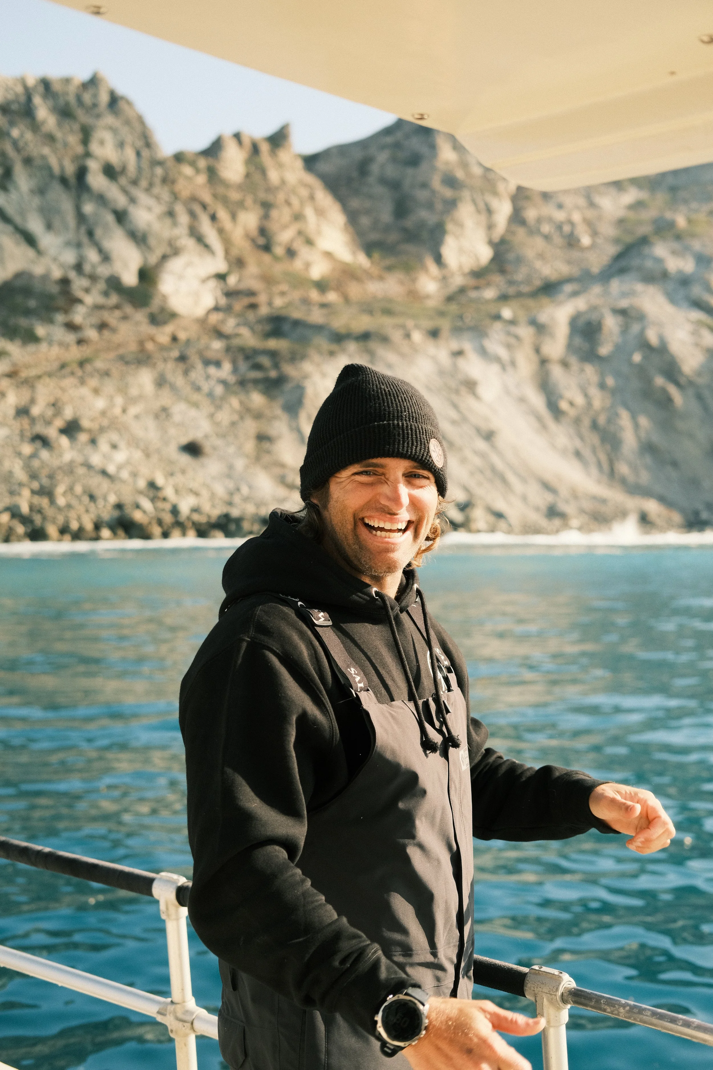A man smiling on a boat with a rocky coastline in the background.
