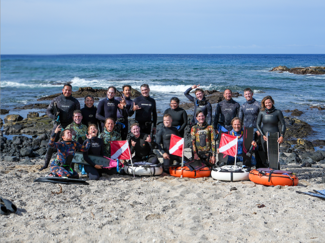 Group of people on a beach with surfboards, wearing wetsuits, smiling, and holding flags.