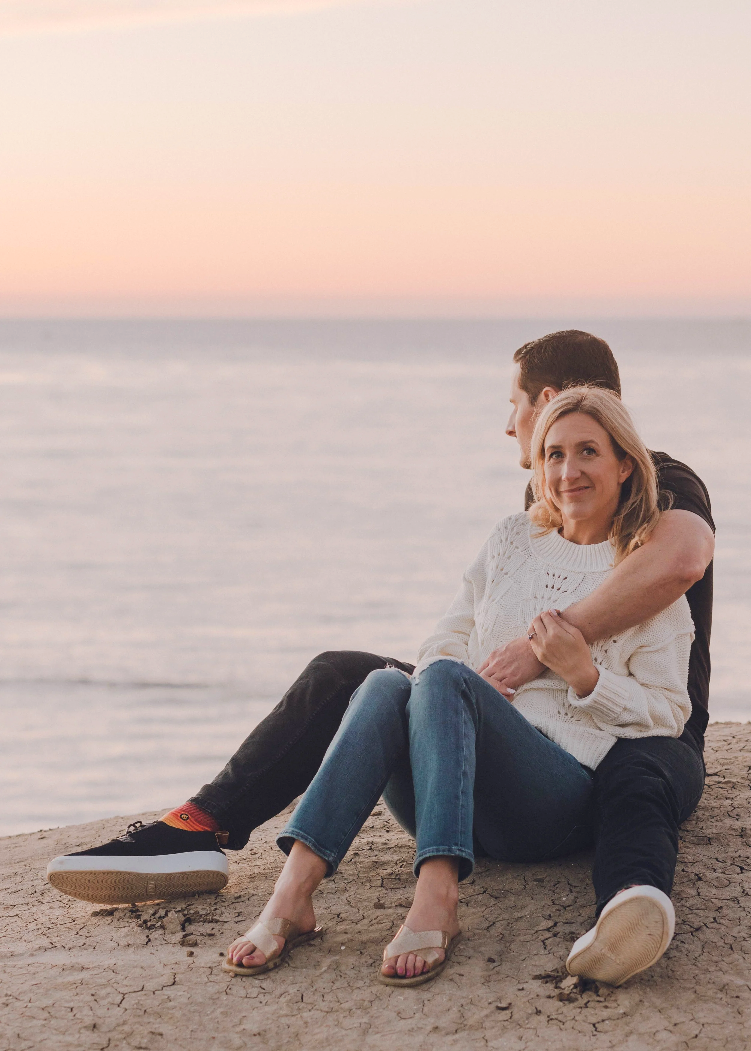 A couple sitting on a cracked dirt surface near the ocean at sunset, with the woman smiling at the camera and the man looking away.
