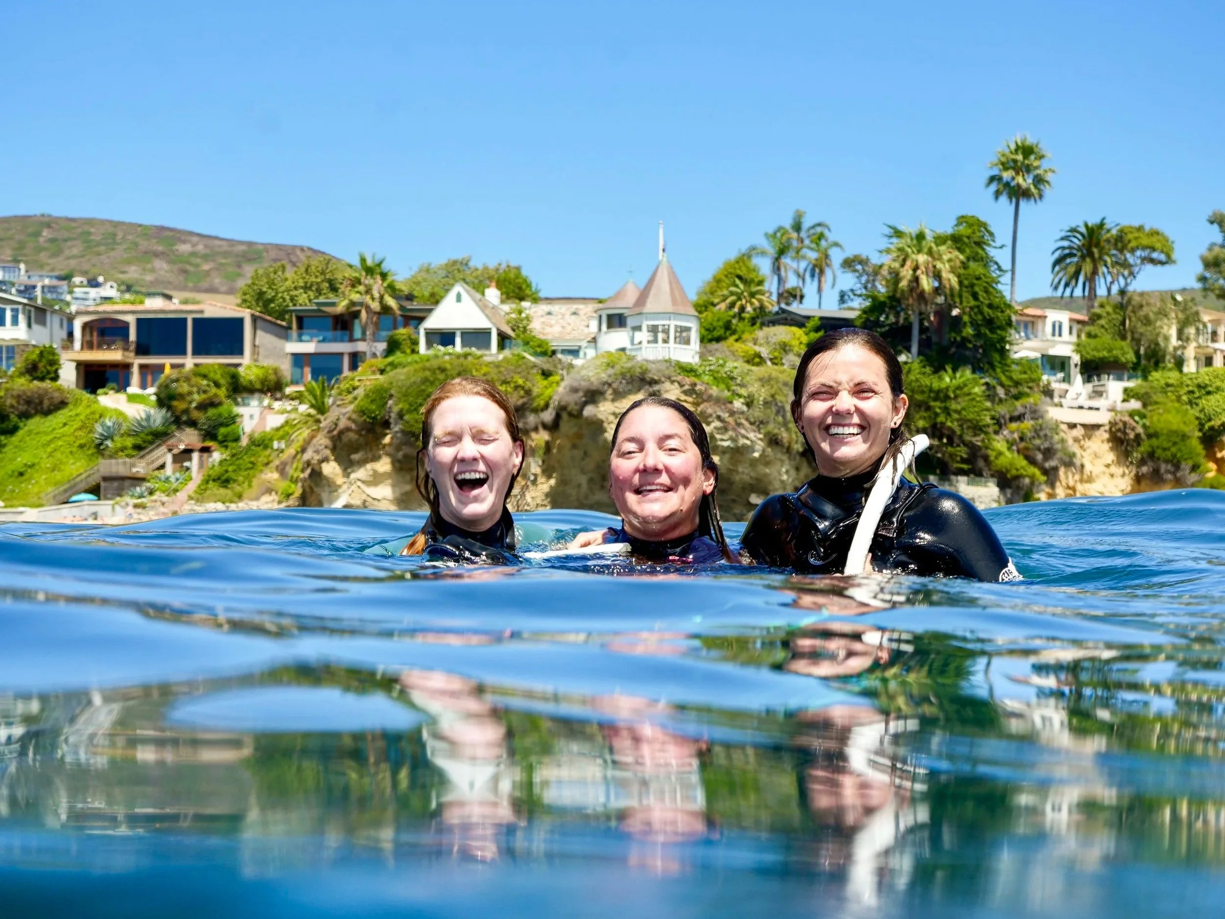 Three women smiling and enjoying themselves in the water with a background of houses, palm trees, and a hill under a clear blue sky.