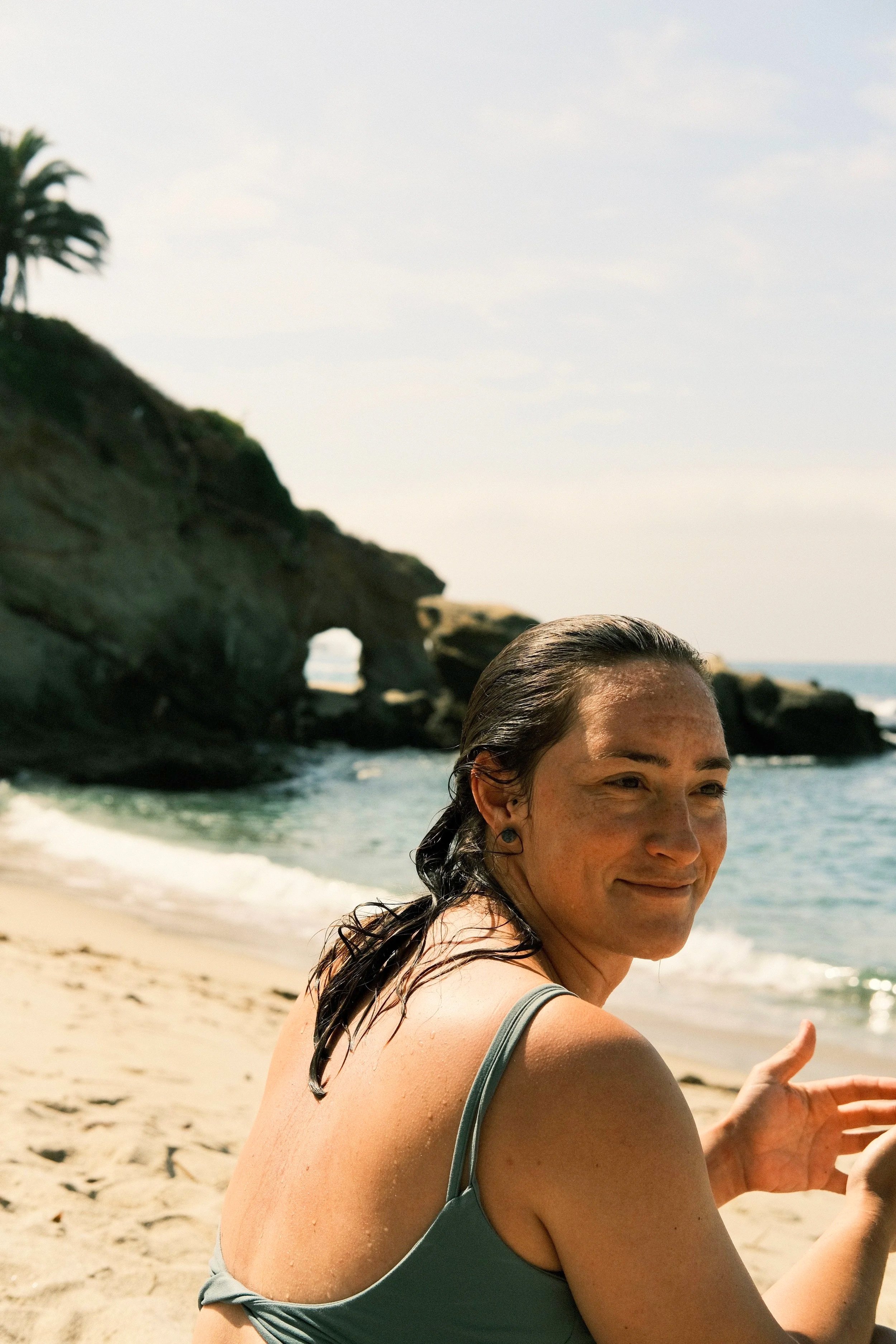 A woman with wet hair sitting on a sandy beach near the ocean, smiling with rocky cliffs and a natural arch in the background.