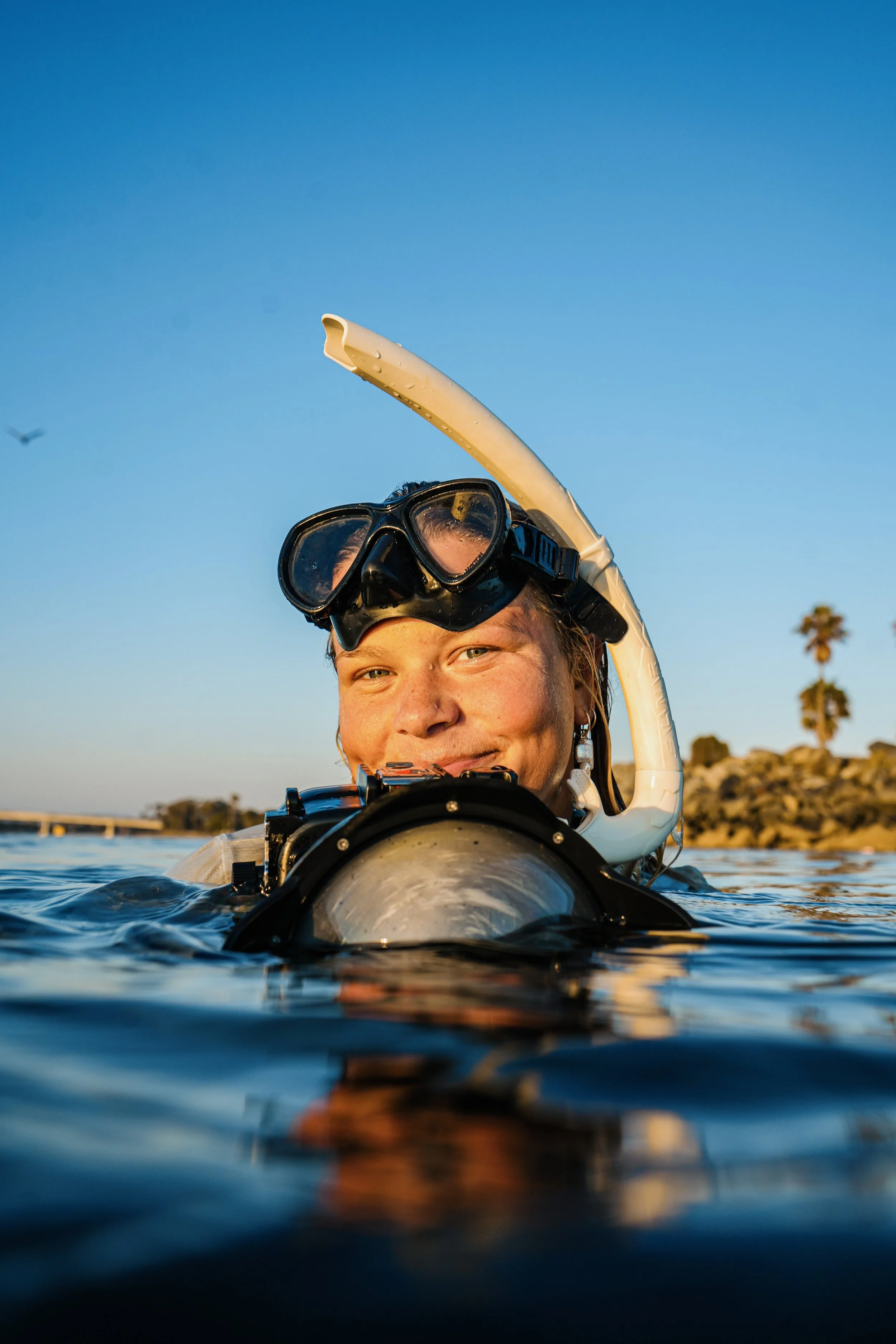 Person wearing a snorkel mask in water, smiling with palm trees and a bridge in the background during sunset.
