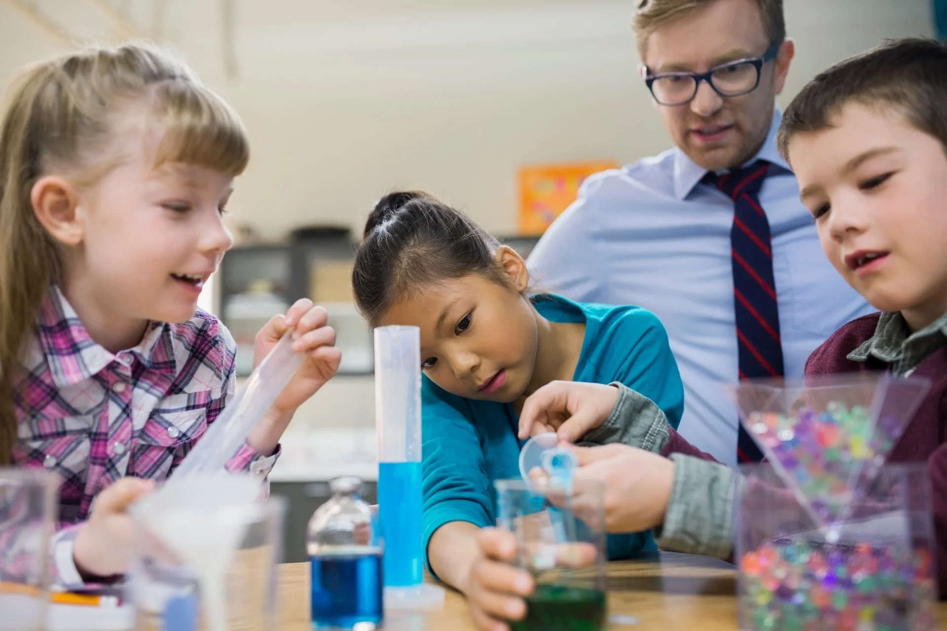 Young students mixing colorful liquids during a chemistry experiment while the teacher supervises.
