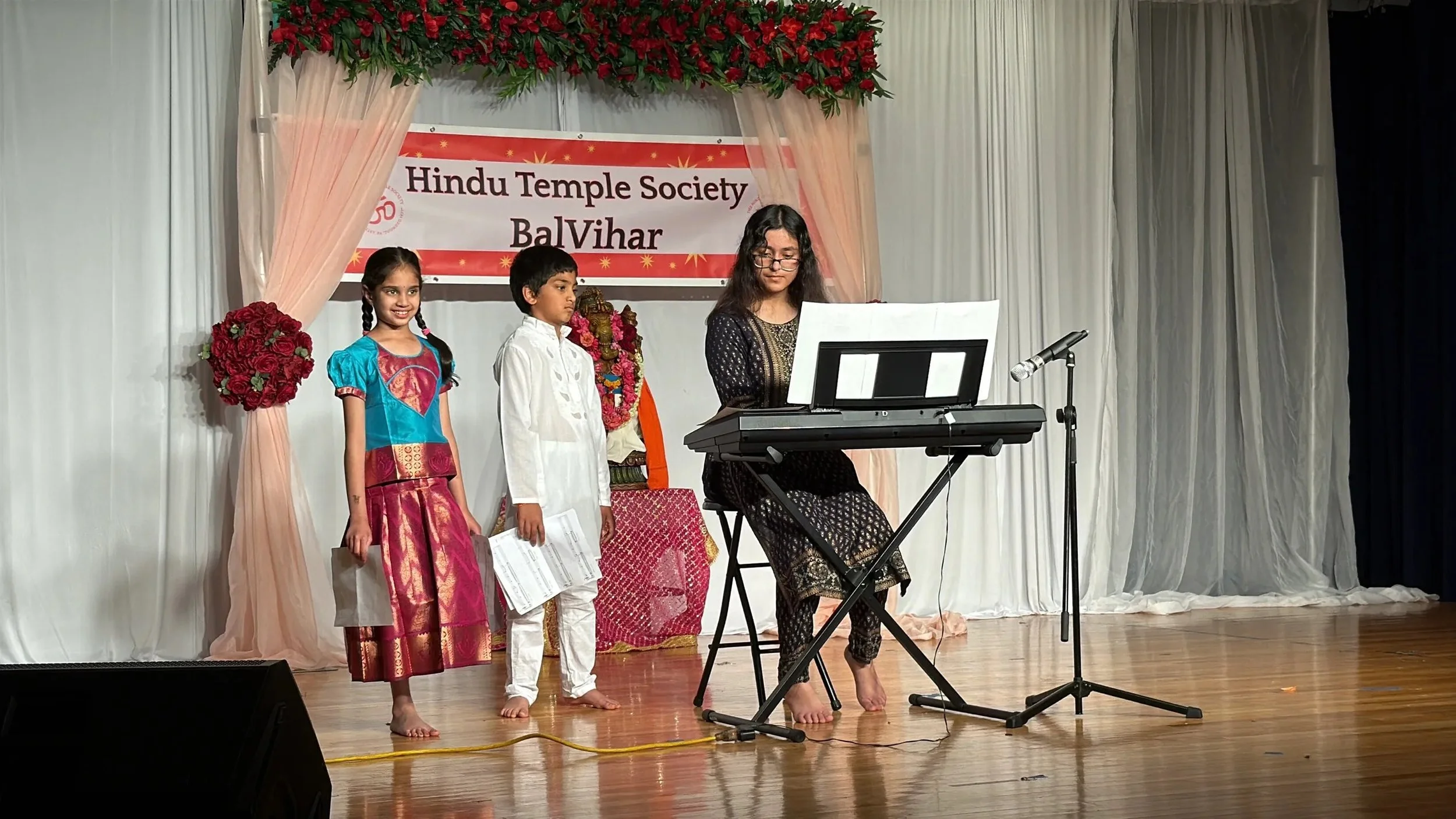A group of three children and one adult woman performing on stage at a Hindu Temple Society event. The woman is playing a keyboard, while the children stand nearby holding papers. The stage is decorated with a banner that reads 'Hindu Temple Society BalVihar' and flowers.