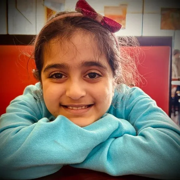 Anora Vikram, Creative Director of Dream2Evolve, with dark hair, wearing a red bow headband and a light blue hoodie, smiling and resting her chin on her arms at a table.