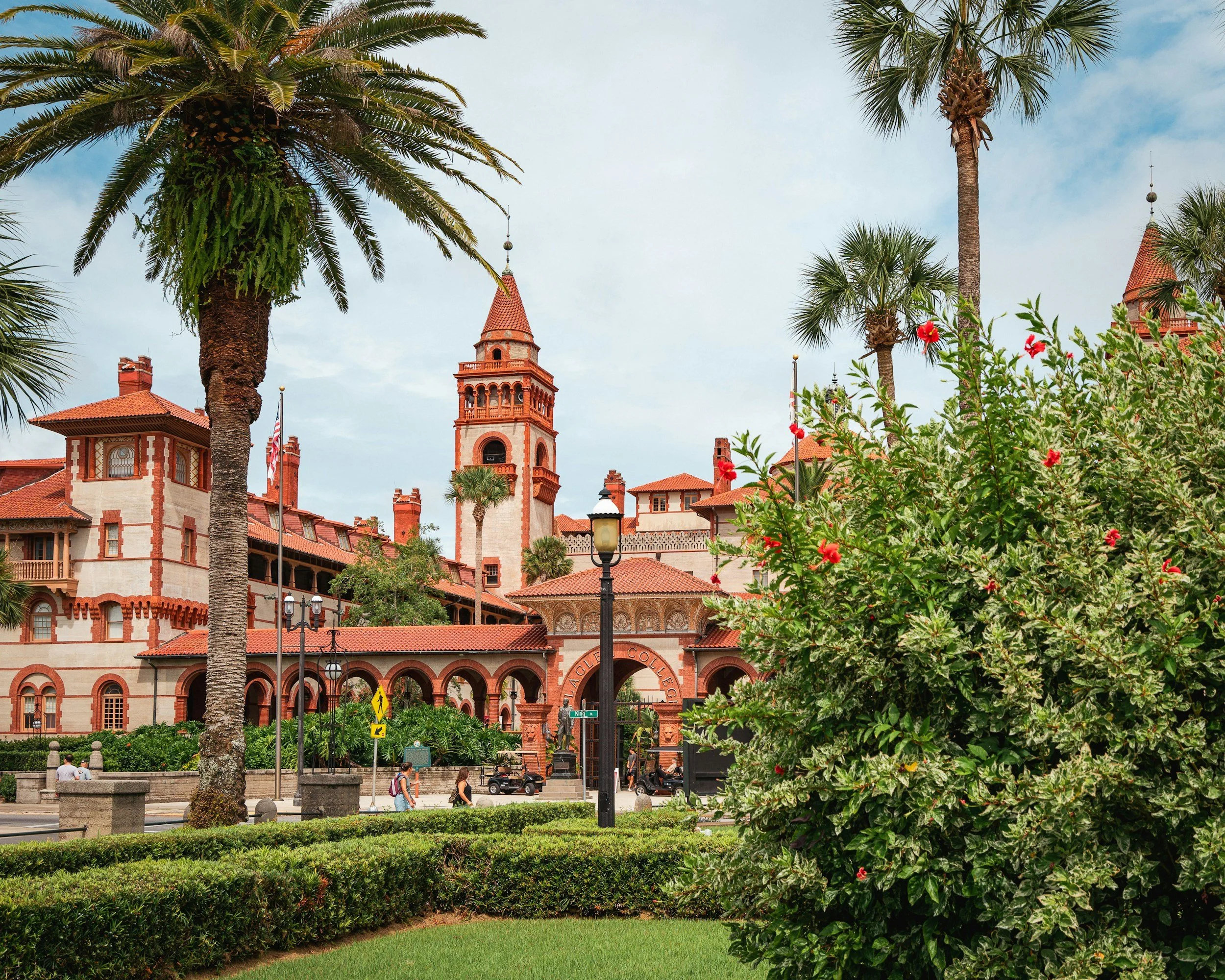 A historic building with a red brick and stucco exterior, surrounded by palm trees, bushes, and greenery. The building features a prominent tower and arched entryway, with people walking nearby.