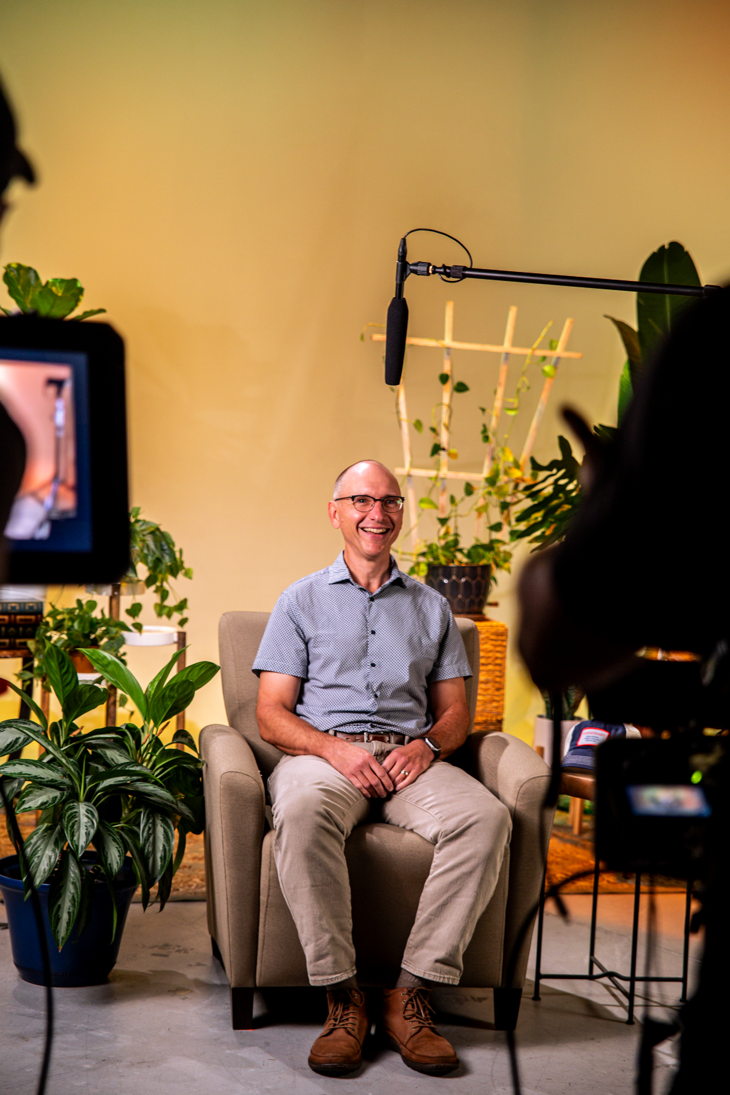 Man sitting in a beige armchair smiling during an interview or video recording in a studio with plants and equipment around him.