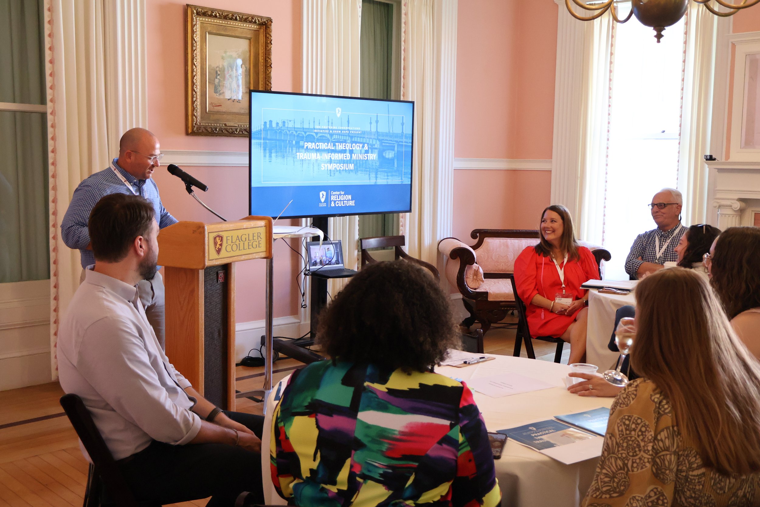 A man in a blue shirt standing at a podium labeled 'Flagler College' giving a presentation to an audience in a room with pink walls and tall windows. A large screen behind him displays a slide titled 'Practical Theology & Trauma-Informed Ministry Symposium.' Several attendees are seated around a table, some smiling and listening attentively.