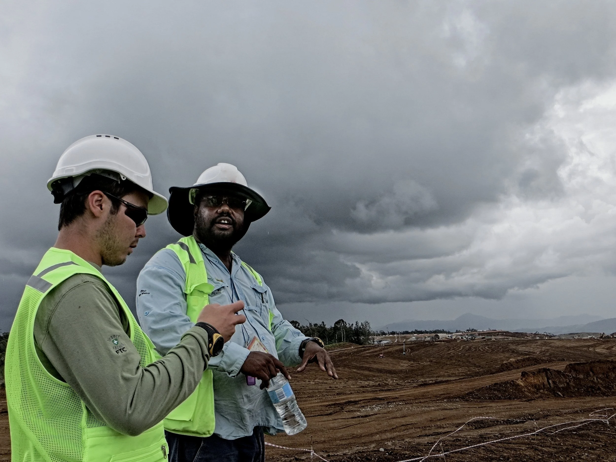 Two construction workers wearing hard hats and high-visibility vests stand against a background of dark, cloudy sky and a construction site with dirt and mountains in the distance.