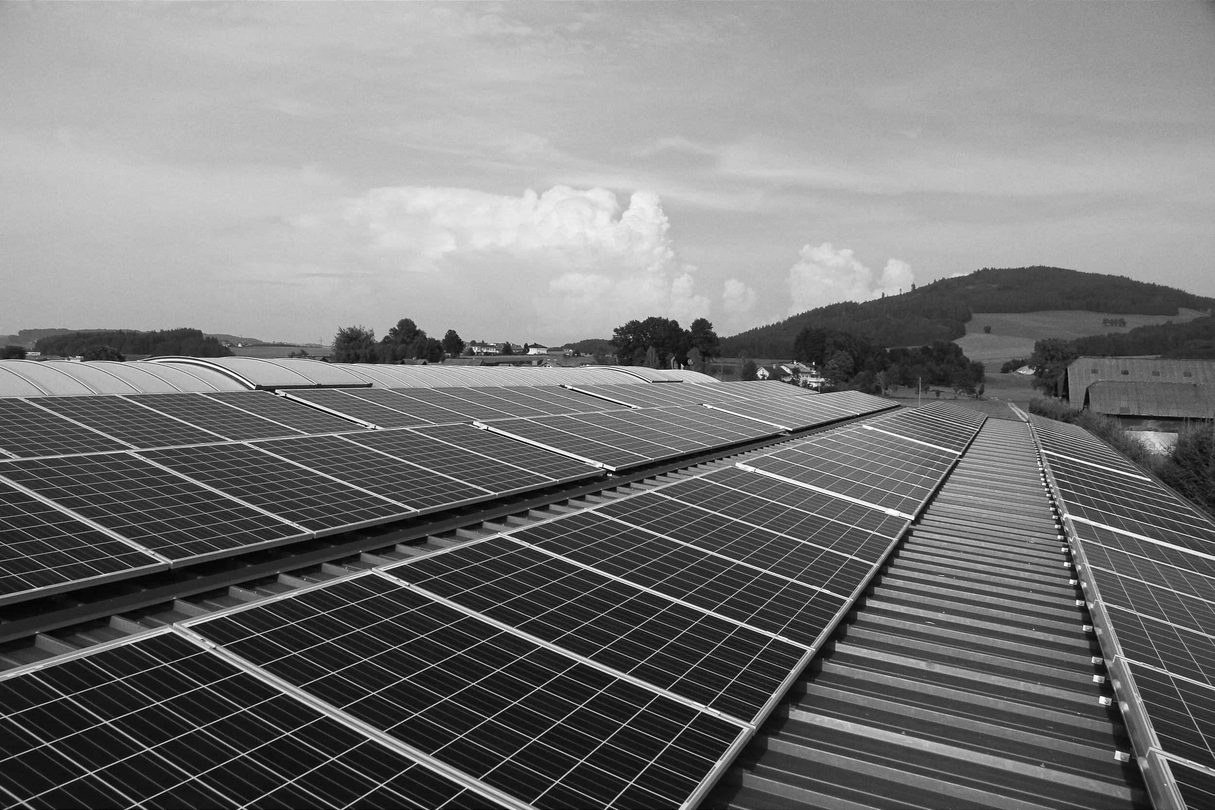 Black and white photo of solar panels installed on a large, curved rooftop, with a rural landscape and hills in the background under a partly cloudy sky.