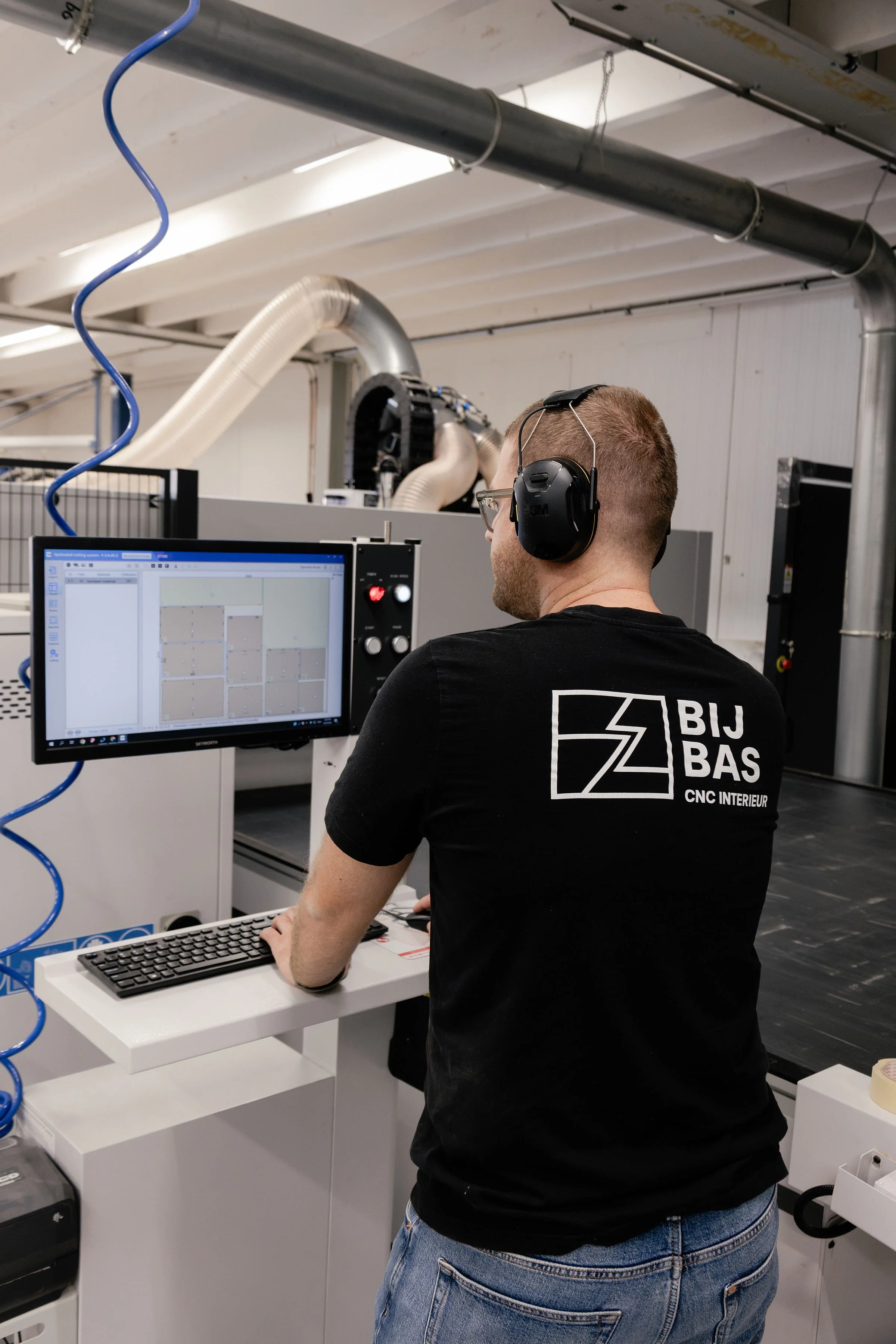 A man wearing headphones and glasses working at a computer in an industrial or workshop environment with visible ducts and piping, wearing a black t-shirt with a logo and text on the back