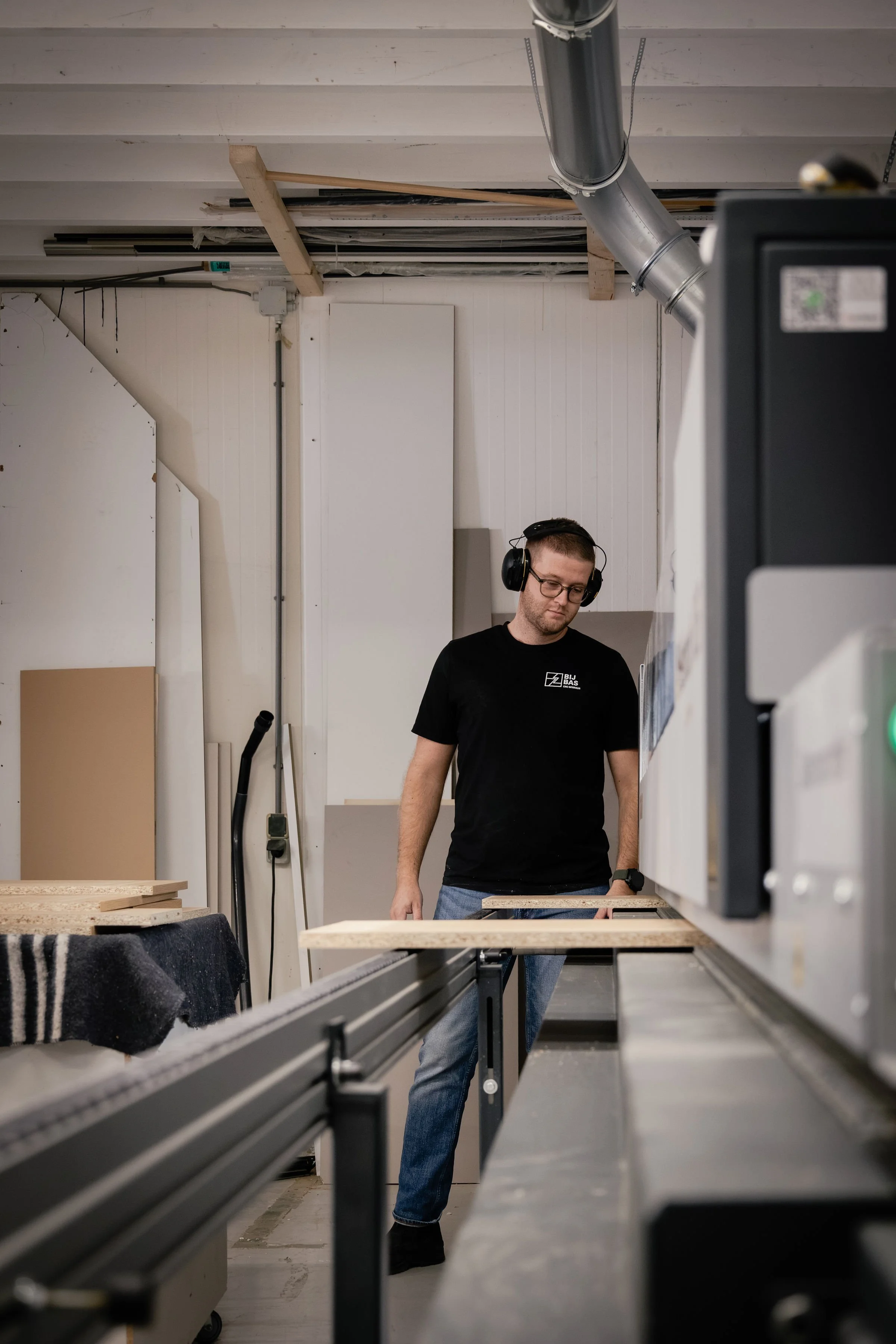 A man working in a workshop wearing safety glasses and headphones, standing near a woodworking table with wood pieces and a large machine.