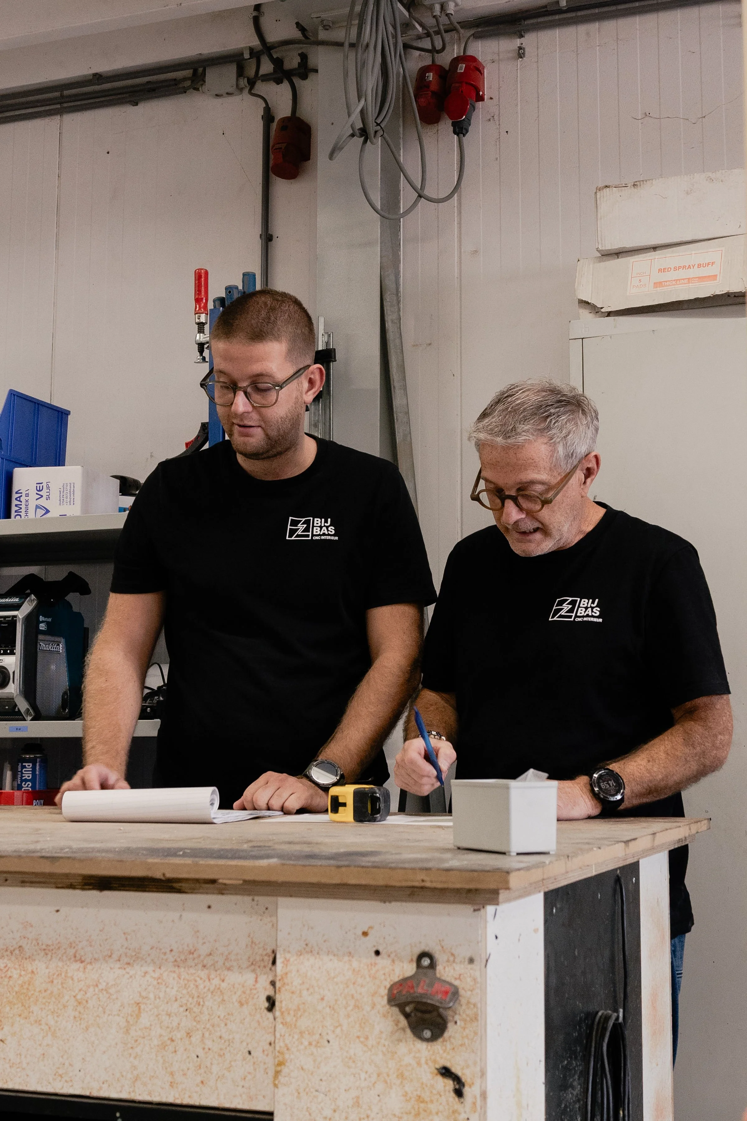 Two men working together in a workshop, wearing black T-shirts with a logo, for a woodworking or construction project, standing over a workbench with tools and papers.
