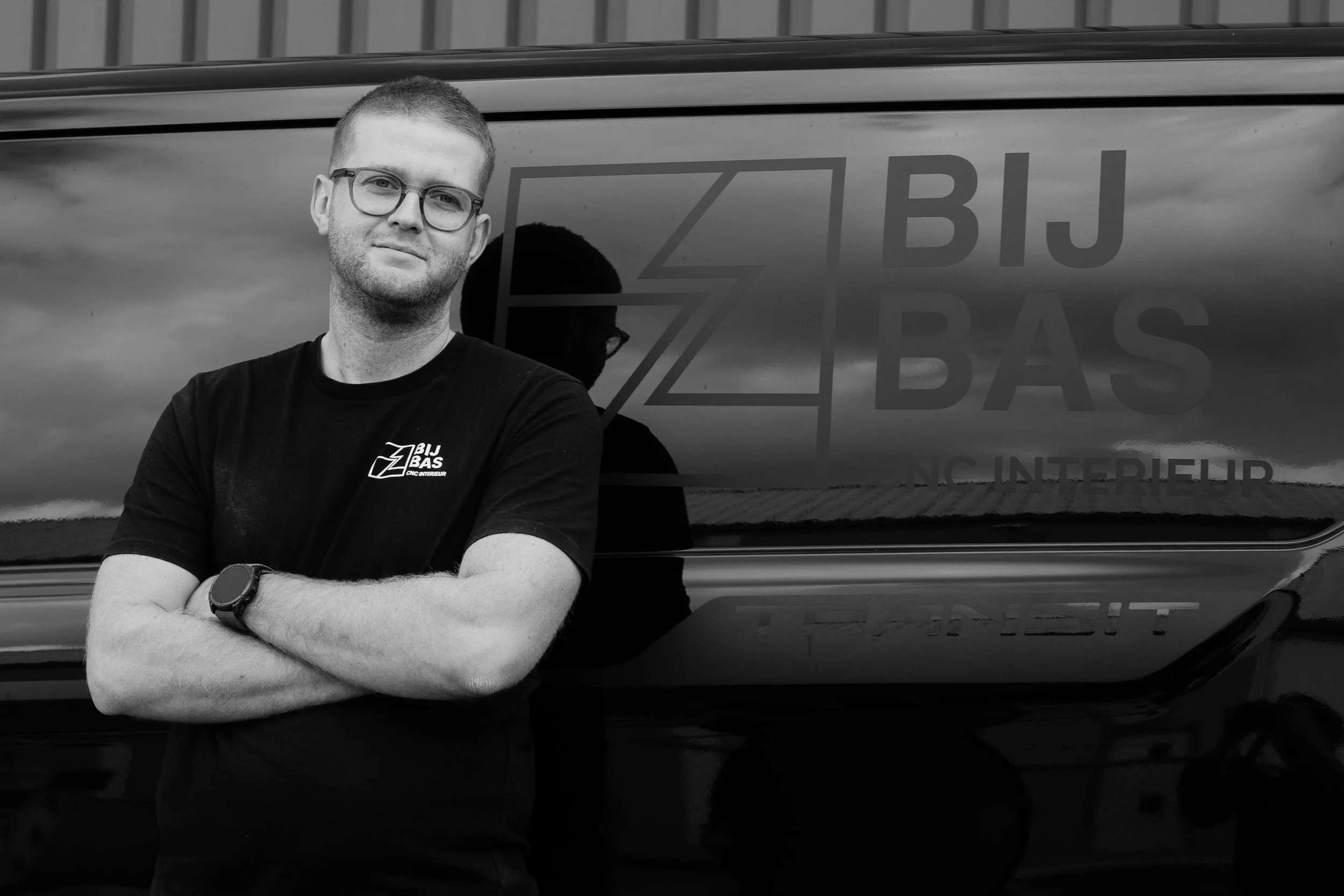 A man wearing glasses and a black T-shirt with a logo stands with his arms crossed in front of a vehicle with a logo that reads 'ZIBJ BAS' and 'NC INTERIEUR'.