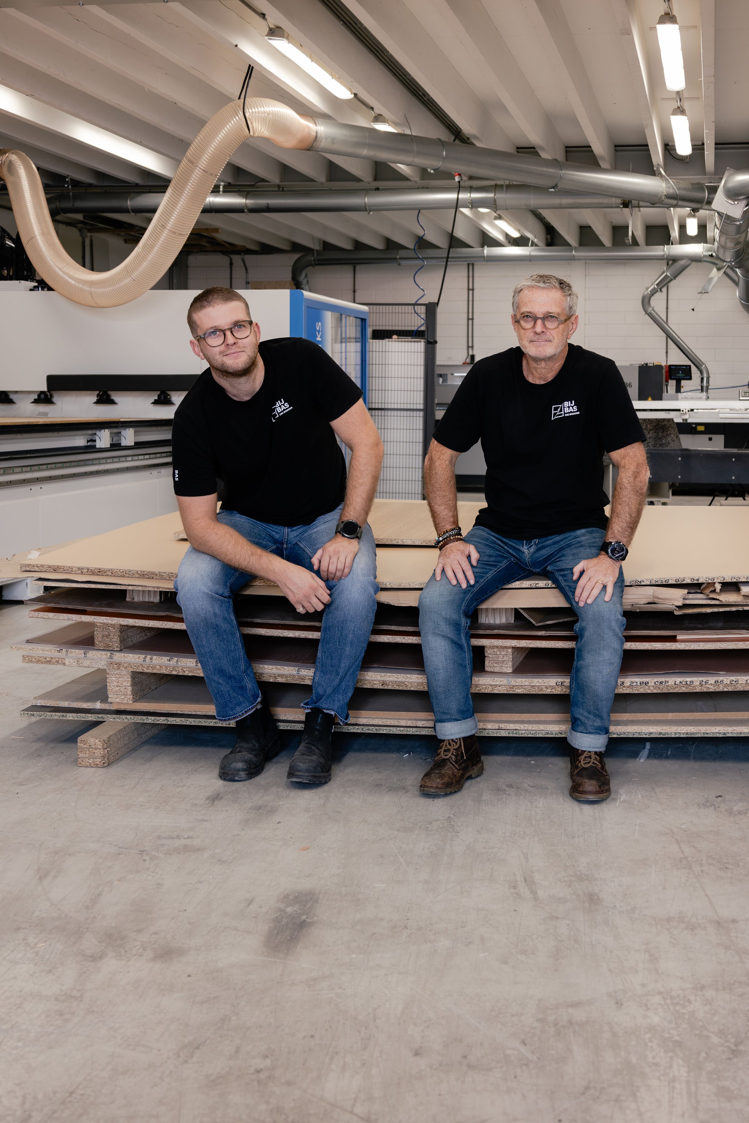 Two men sitting on a wooden pallet in an industrial workshop. They are both wearing black T-shirts with a logo, jeans, and glasses. The background includes industrial pipes, machinery, and a white brick wall.