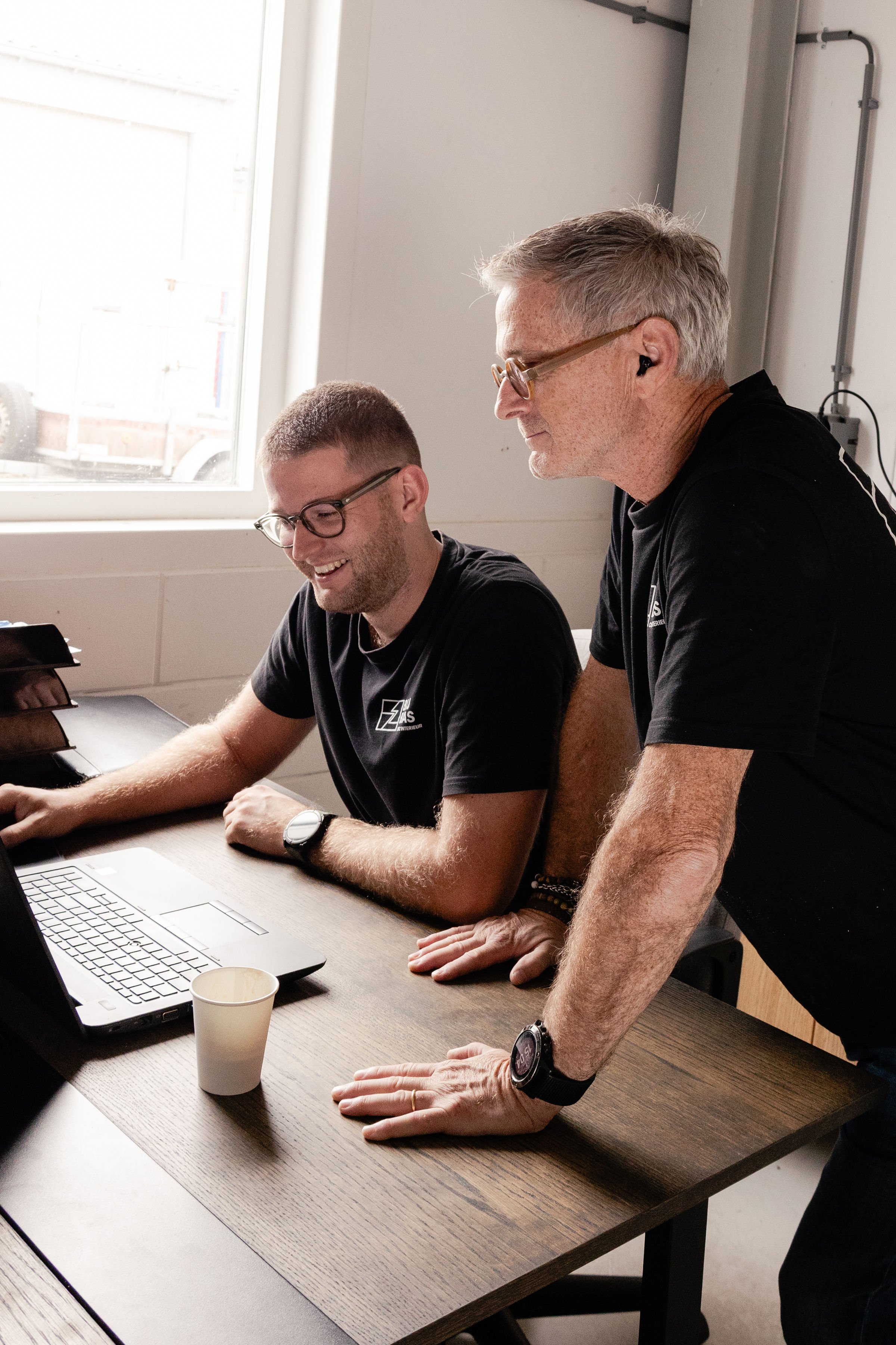 Two men seated at a table, working together on a laptop, with one man standing and looking at the screen. They are smiling and seem engaged in a discussion. The table has a laptop, a white cup, and some papers. The setting appears to be an office or meeting room with a window in the background.