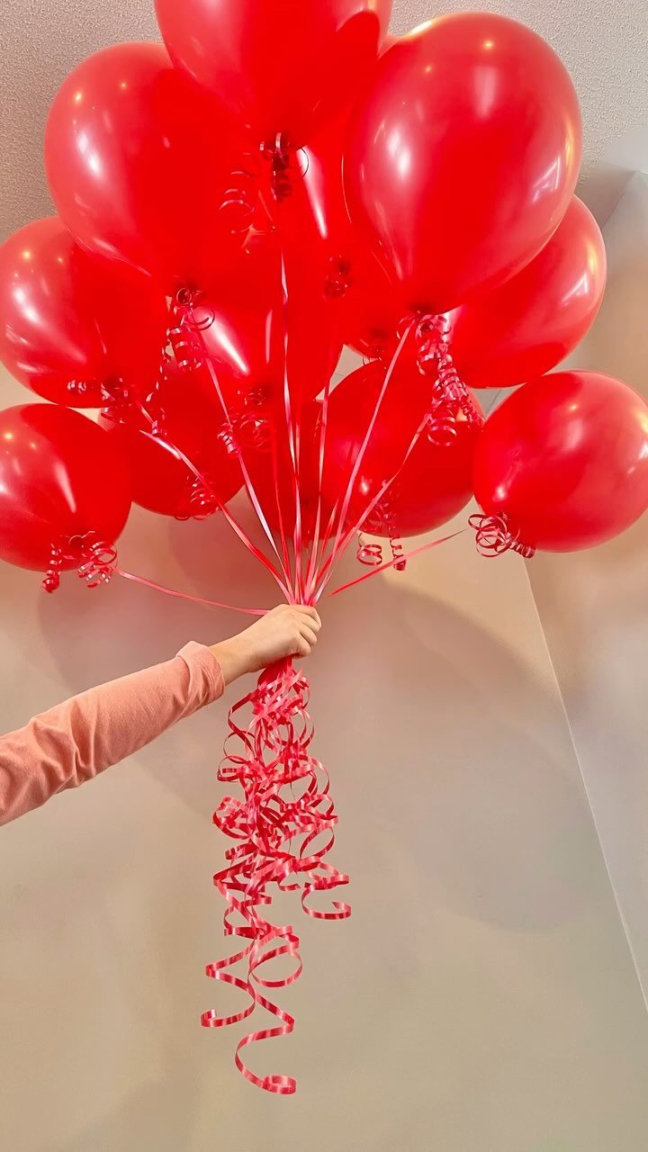 A person holding a large bunch of red balloons with curling ribbons hanging down, set against an indoor wall.