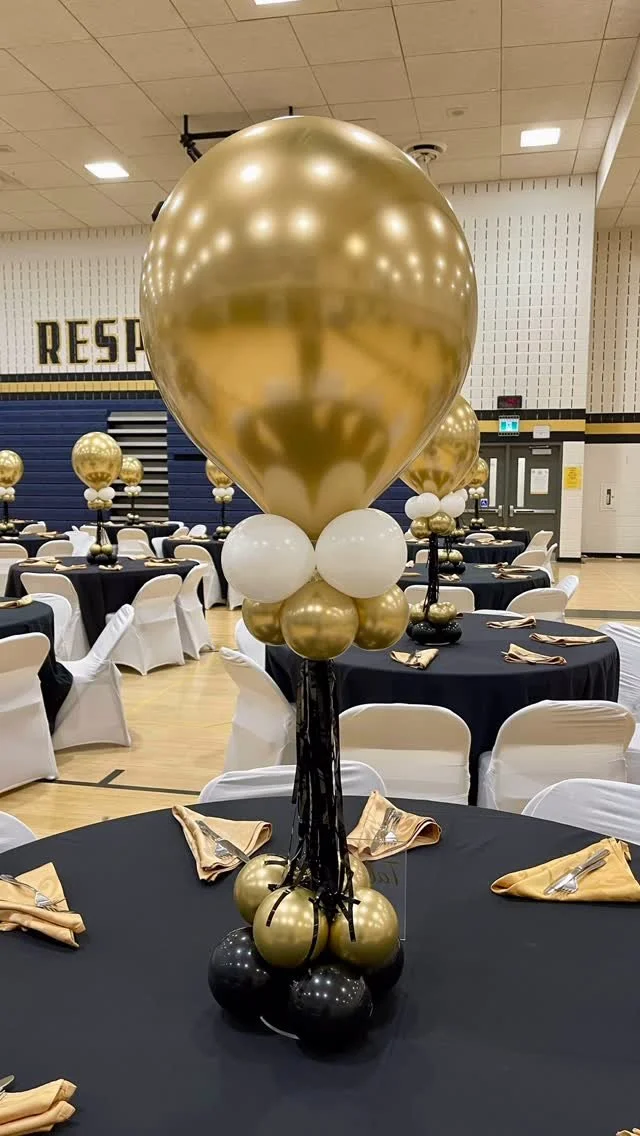 Gold, black, and white balloon centerpiece on a banquet table with black tablecloths and gold napkins in a decorated event hall.