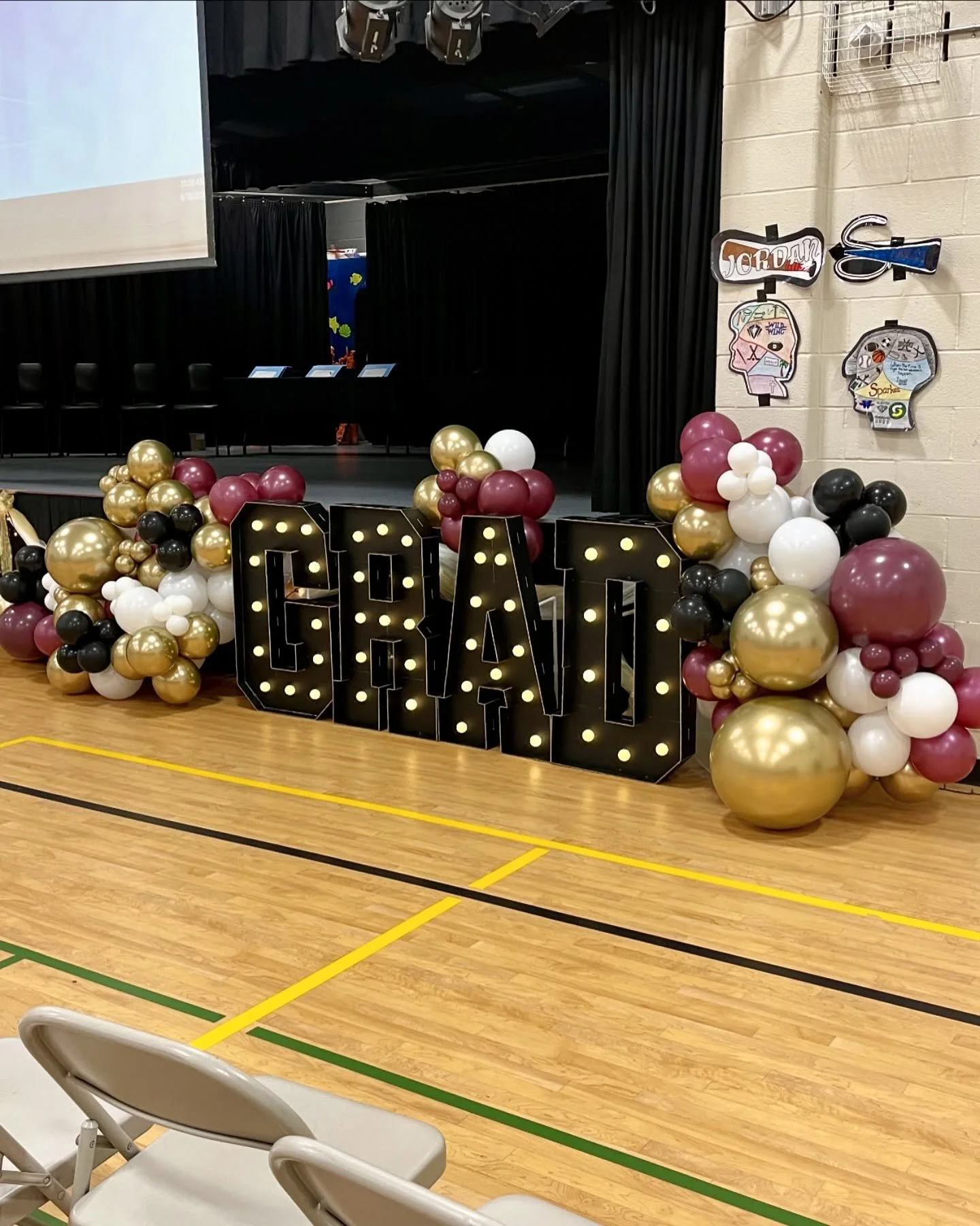 Decorative stage setup with large black marquee letters spelling 'GRAD' illuminated by small light bulbs, surrounded by pink, white, black, and gold balloons arranged in clusters on a gymnasium floor.