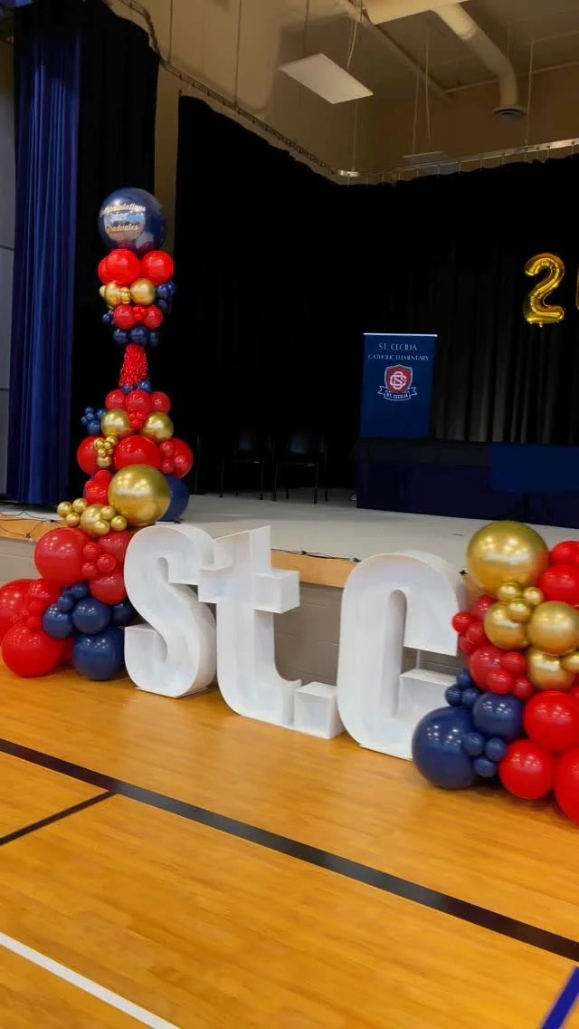 Decorations for a celebration at a school gymnasium, featuring large white letters spelling 'STOP' and balloon arrangements in red, blue, and gold. A stage with a blue banner and a large gold number 2 balloon are visible.