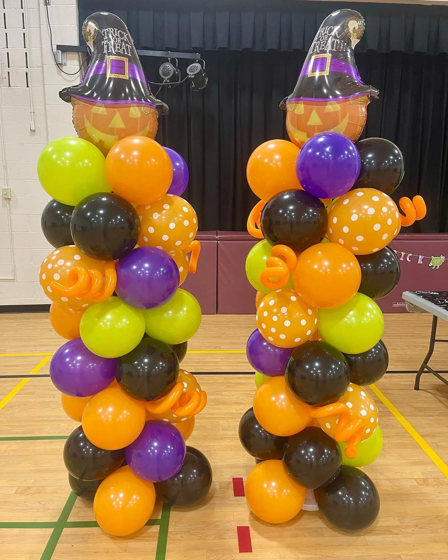 Two large Halloween balloon columns featuring a witch hat balloon topper, orange, black, purple, and lime green balloons, some with polka dots, and orange curly ribbon accents, set up in a gymnasium with a black stage curtain.