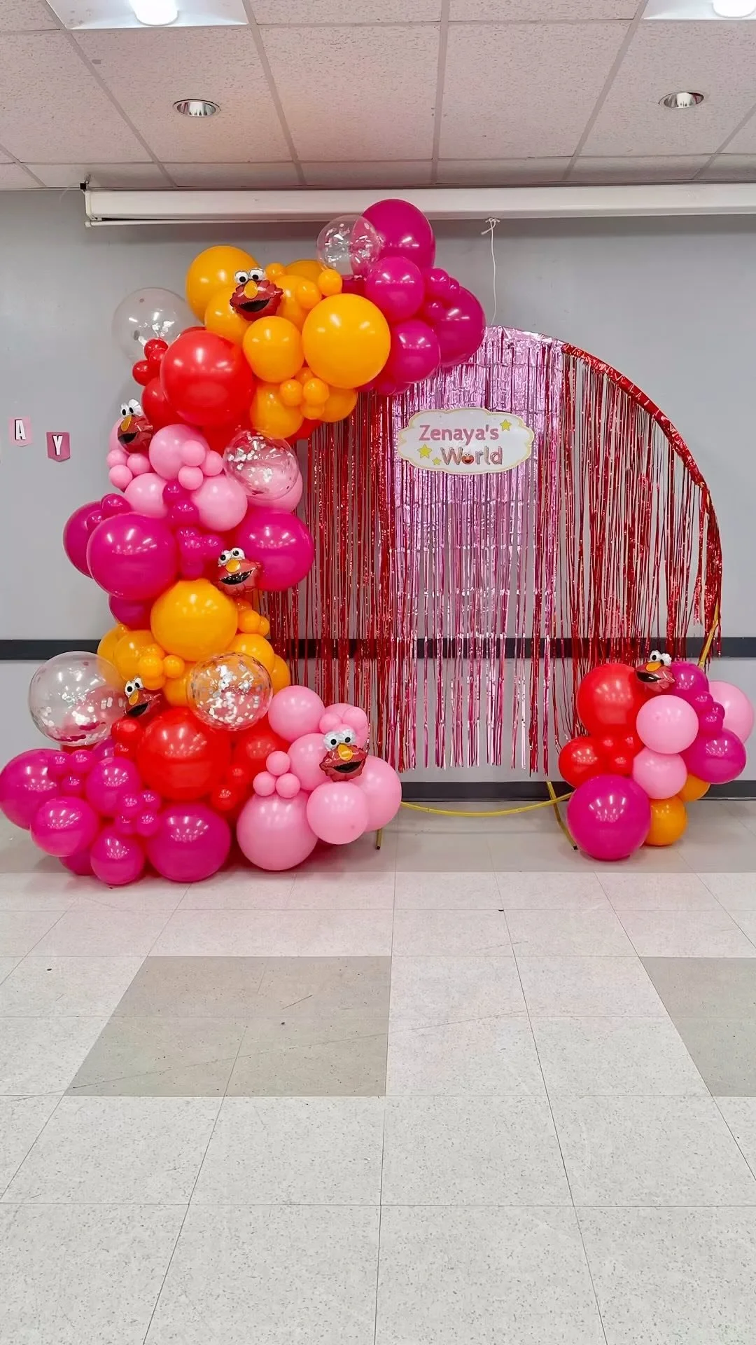 Colorful balloon arch and backdrop decorated with pink, red, orange, and clear balloons, some with cartoon face masks, and a sign that reads 'Zenaya's World' at a children's birthday party or celebration.