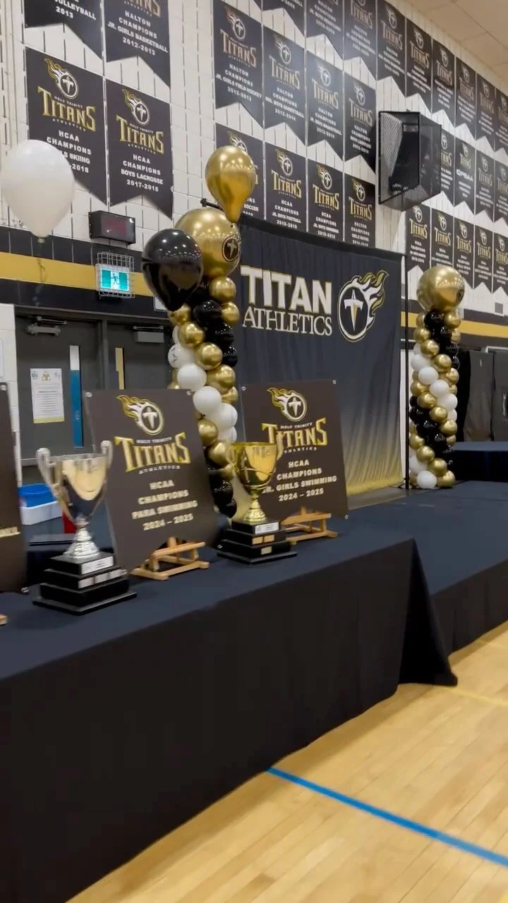 Awards display with trophies, black and gold balloons, and banners celebrating the Titans athletics championships in men's and women's swimming, 2014-2025, in a gymnasium.