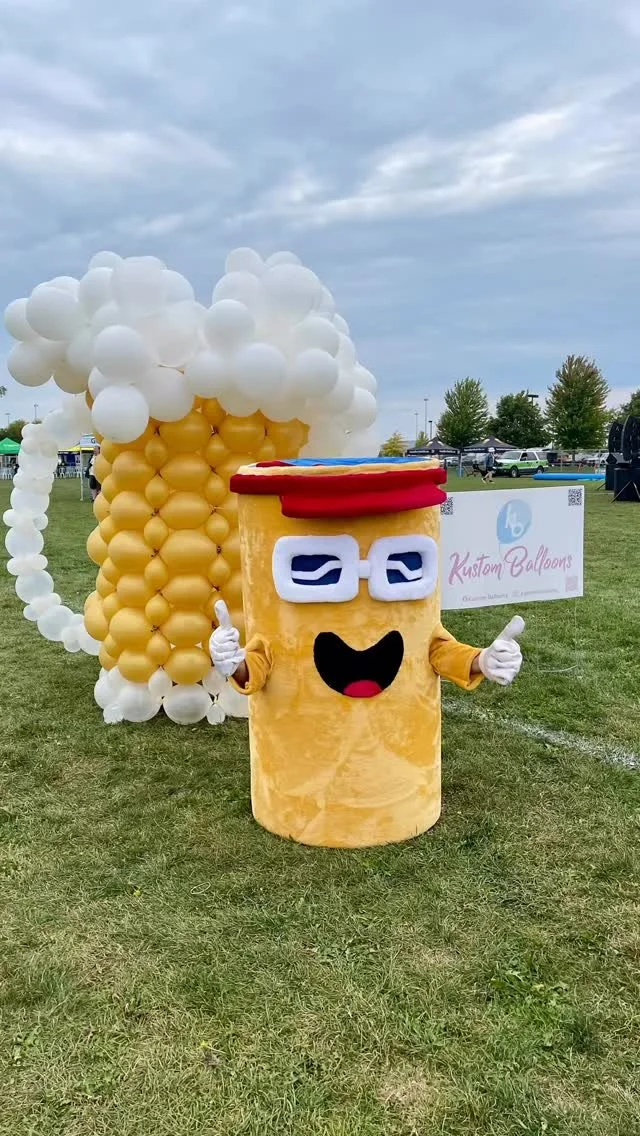 A person dressed up in a balloon costume resembling a coffee cup with glasses and a red hat, giving a thumbs-up gesture at an outdoor event, with a large cluster of white and yellow balloons in the background.