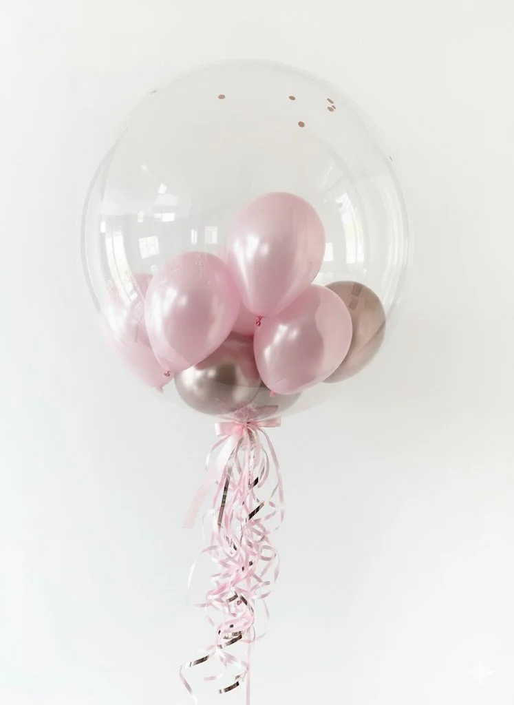 A transparent balloon filled with pink and metallic silver smaller balloons, with pink curly ribbon decorations hanging from the bottom.