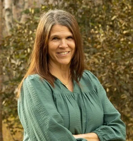A woman with long brown hair smiling outdoors, wearing a teal blouse, with trees and autumn foliage in the background.