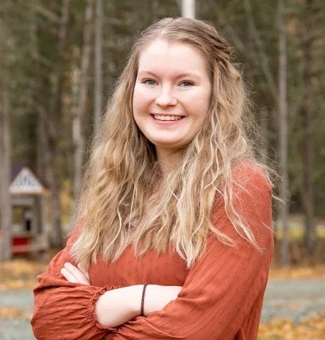 Young woman with long blonde curly hair smiling outdoors with trees and a small building in background.