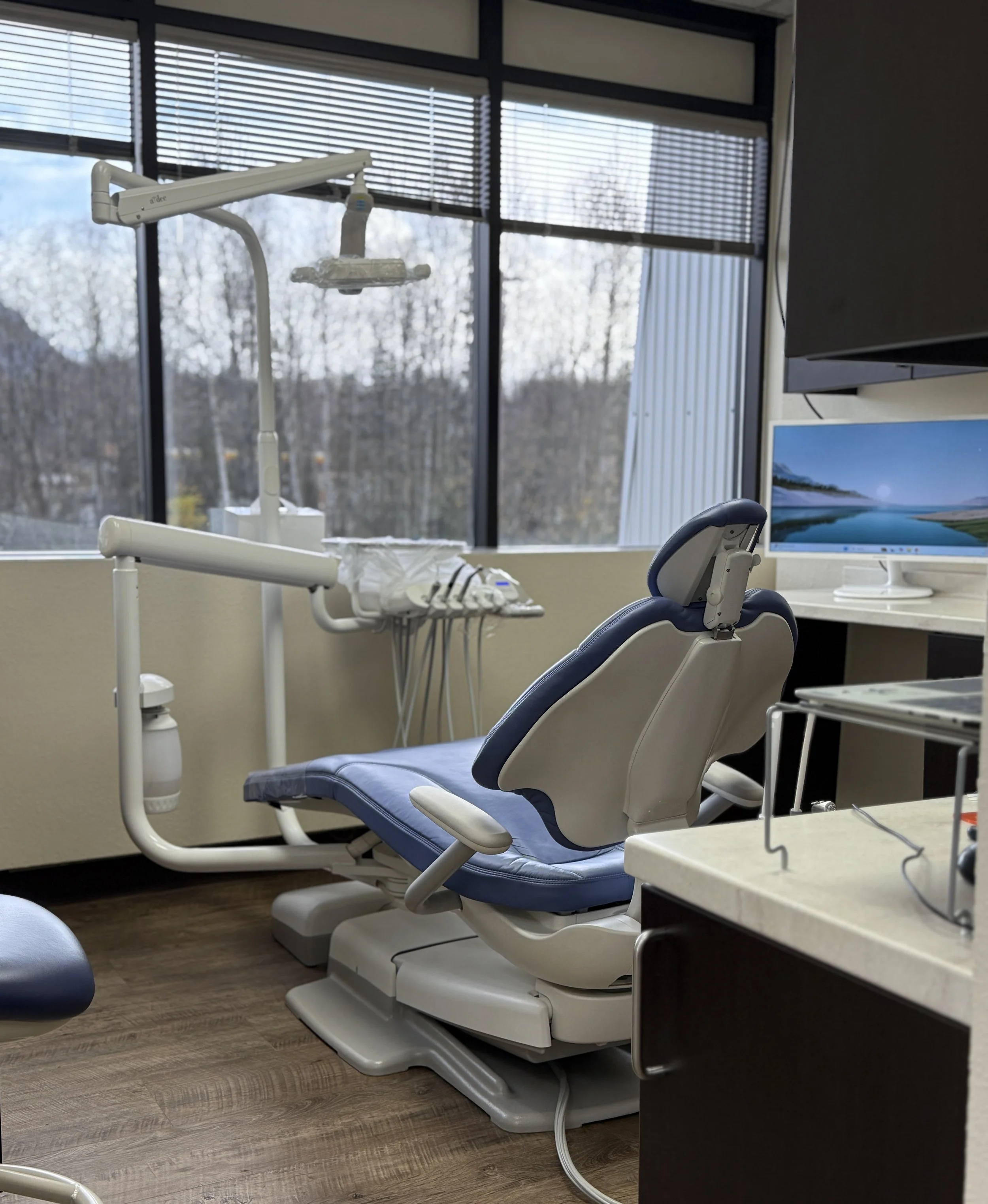 Empty dental examination chair in a dentist's office with dental equipment nearby and a large window in the background.