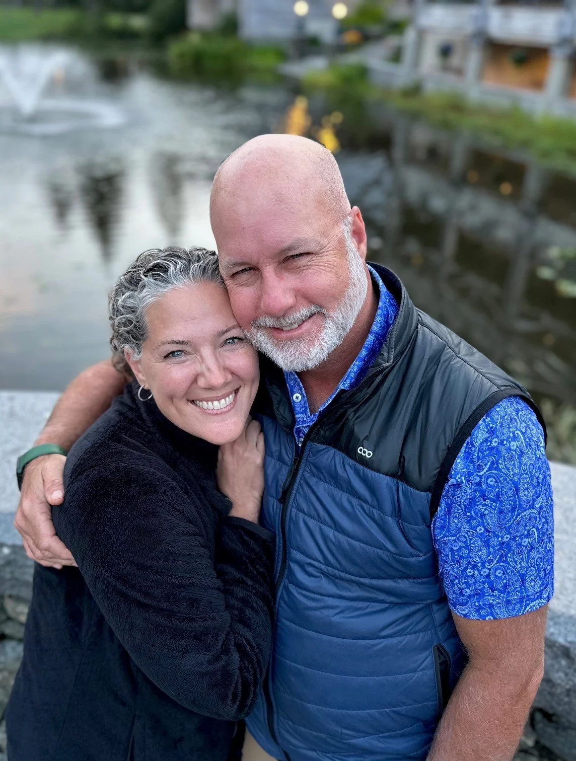 A smiling mature couple hugging outdoors near a body of water, with houses blurred in the background.