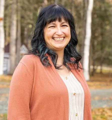 Woman with dark hair and a smile, wearing a peach-colored jacket over a white striped shirt, standing outdoors in a park with trees and fallen leaves.