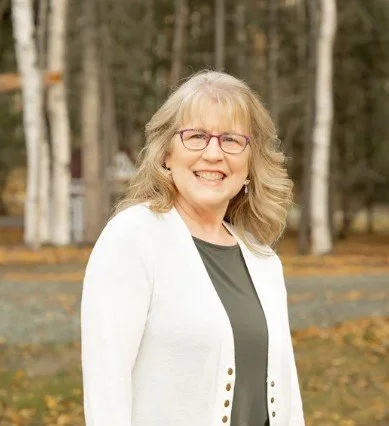 Christy L. with glasses and blonde hair outdoors, wearing a white blazer over a gray top with a background of trees and fallen leaves.