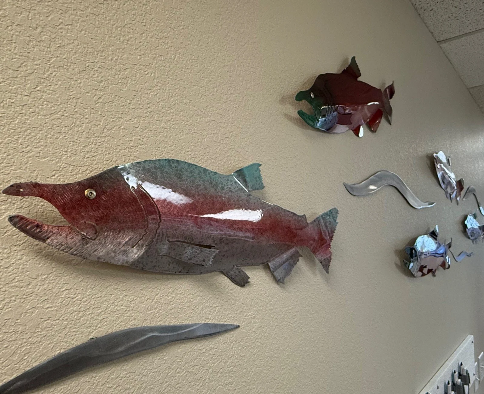 Metal wall art of various fish mounted on a beige wall, including a large fish with a red and gray coloration, and smaller fish sculptures.