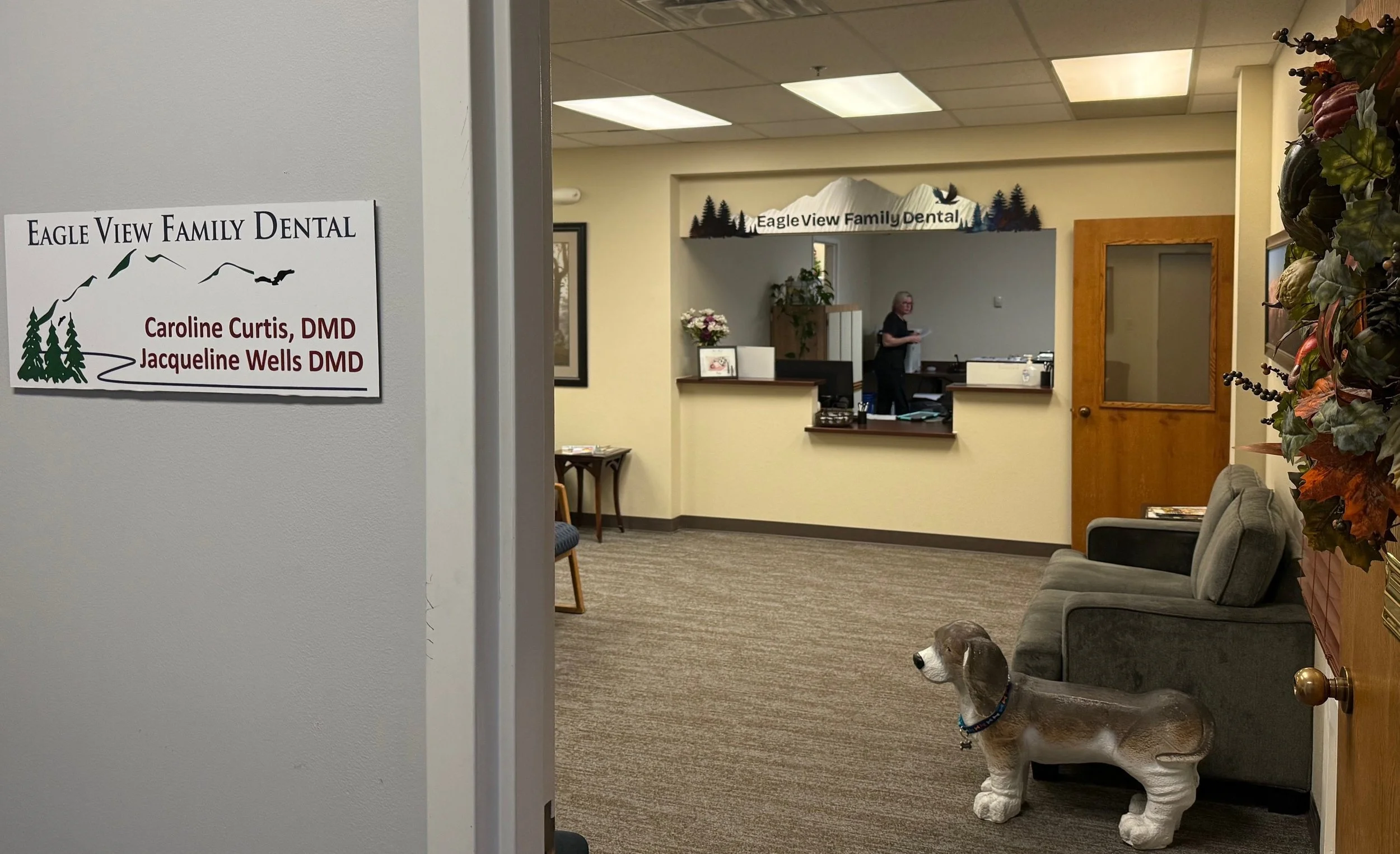 Reception area of Eagle View Family Dental with a sign for Dr. Caroline Curtis DMD and Dr. Jacqueline Wells DMD on the left, a waiting area with chairs and a dog-shaped statue, and a staff member at the reception desk in the background.