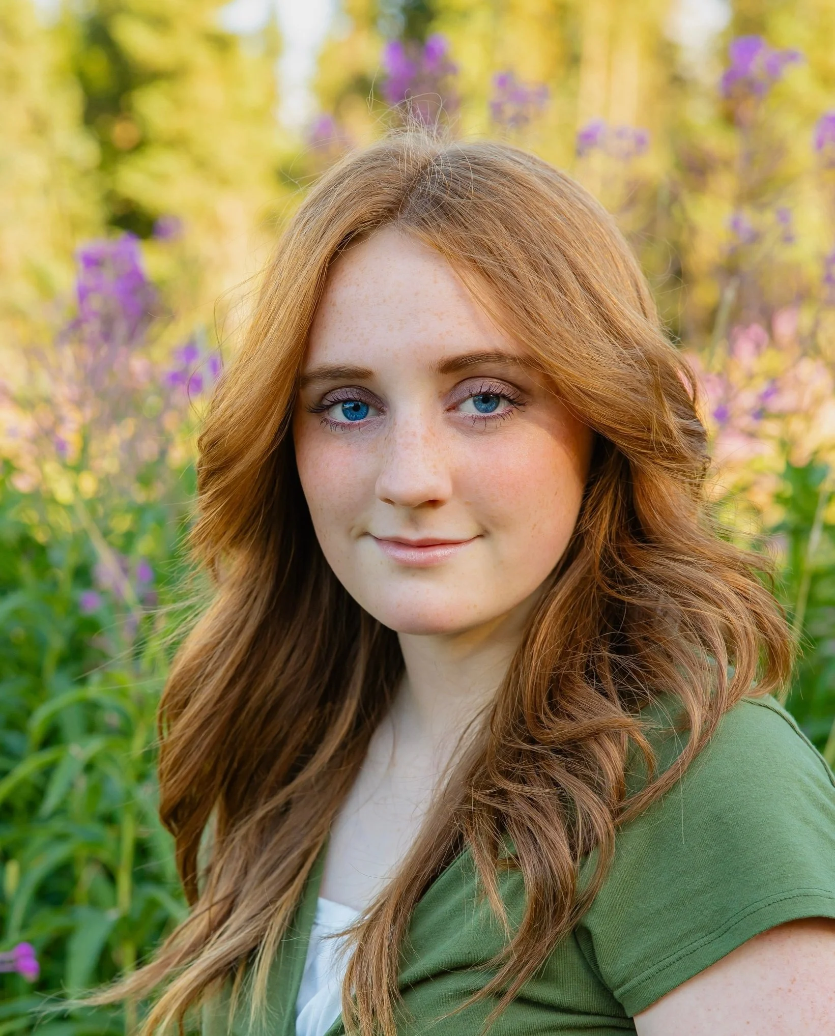A young woman with long red hair and blue eyes standing outdoors in a garden with purple flowers and trees.
