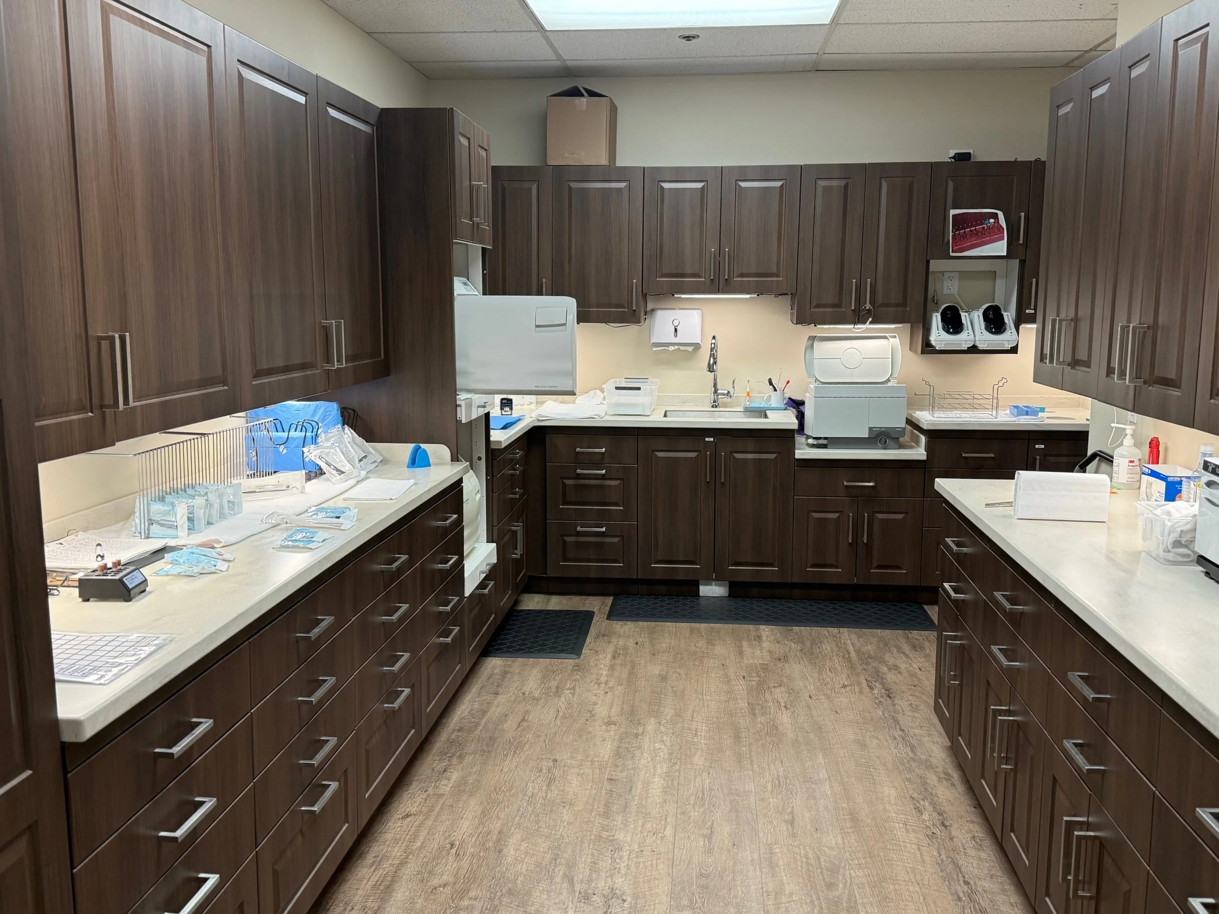 A hospital or medical laboratory kitchen with dark brown cabinets and white countertops, containing medical supplies and equipment, including a sterilizer, disinfectants, and sample trays.