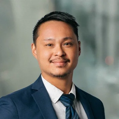 Danny with short, black hair, a slight mustache and beard, wearing a blue suit and blue tie, smiles at the camera.