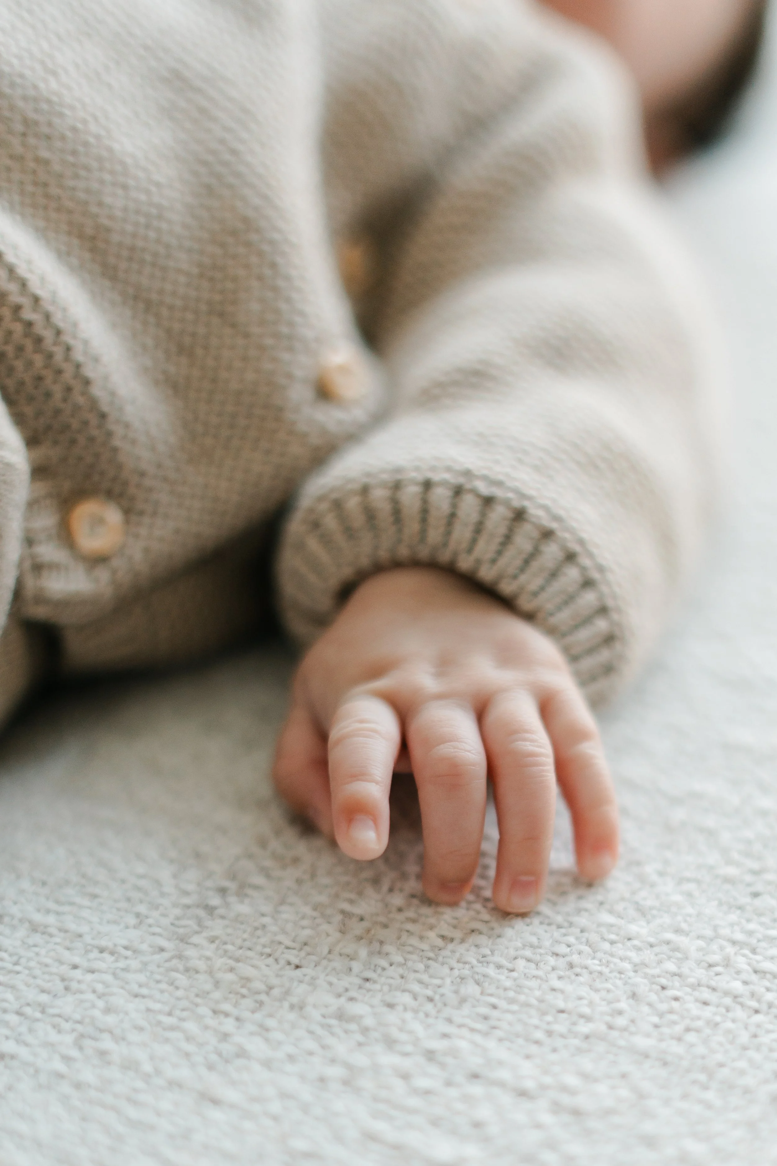 Close-up van een kinderhand rustend op een beige tapijt, gekleed in een grijs gebreid truitje met knopen.