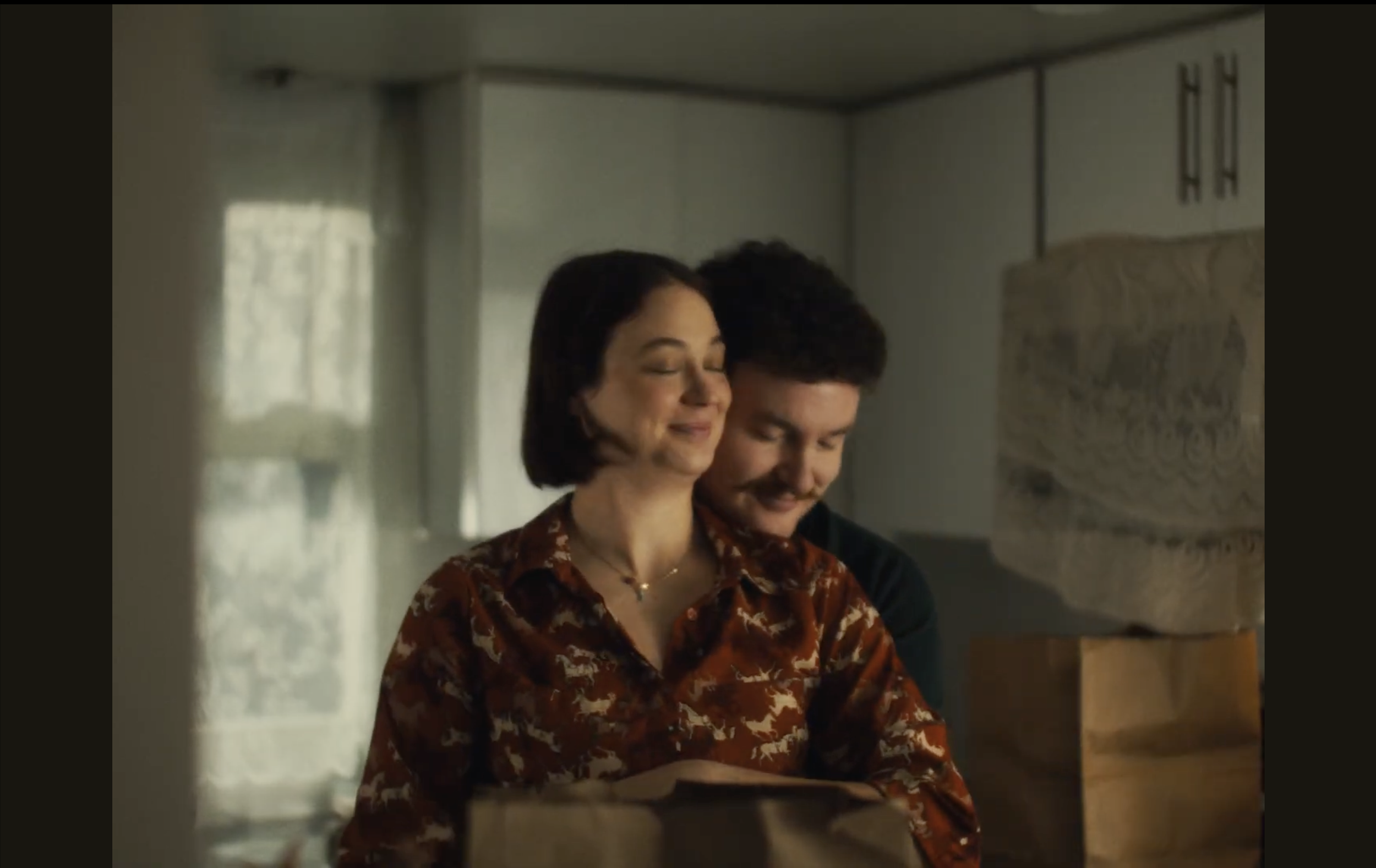 A woman in a patterned brown blouse smiling with closed eyes, standing close to a man in a dark shirt, both smiling softly, inside a room with a painting on the wall and a window with curtains.