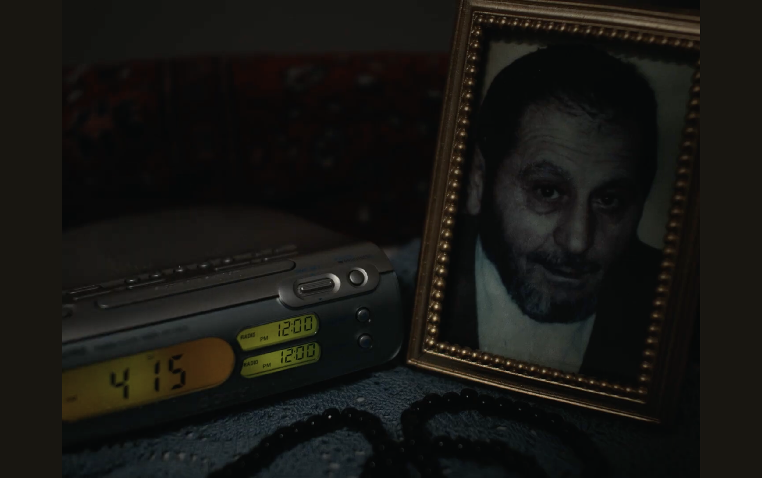 A framed black and white portrait of a man with dark hair and a beard, a digital alarm clock showing 12:00, and a yellow digital clock showing 4:15. The items are on a dark surface.