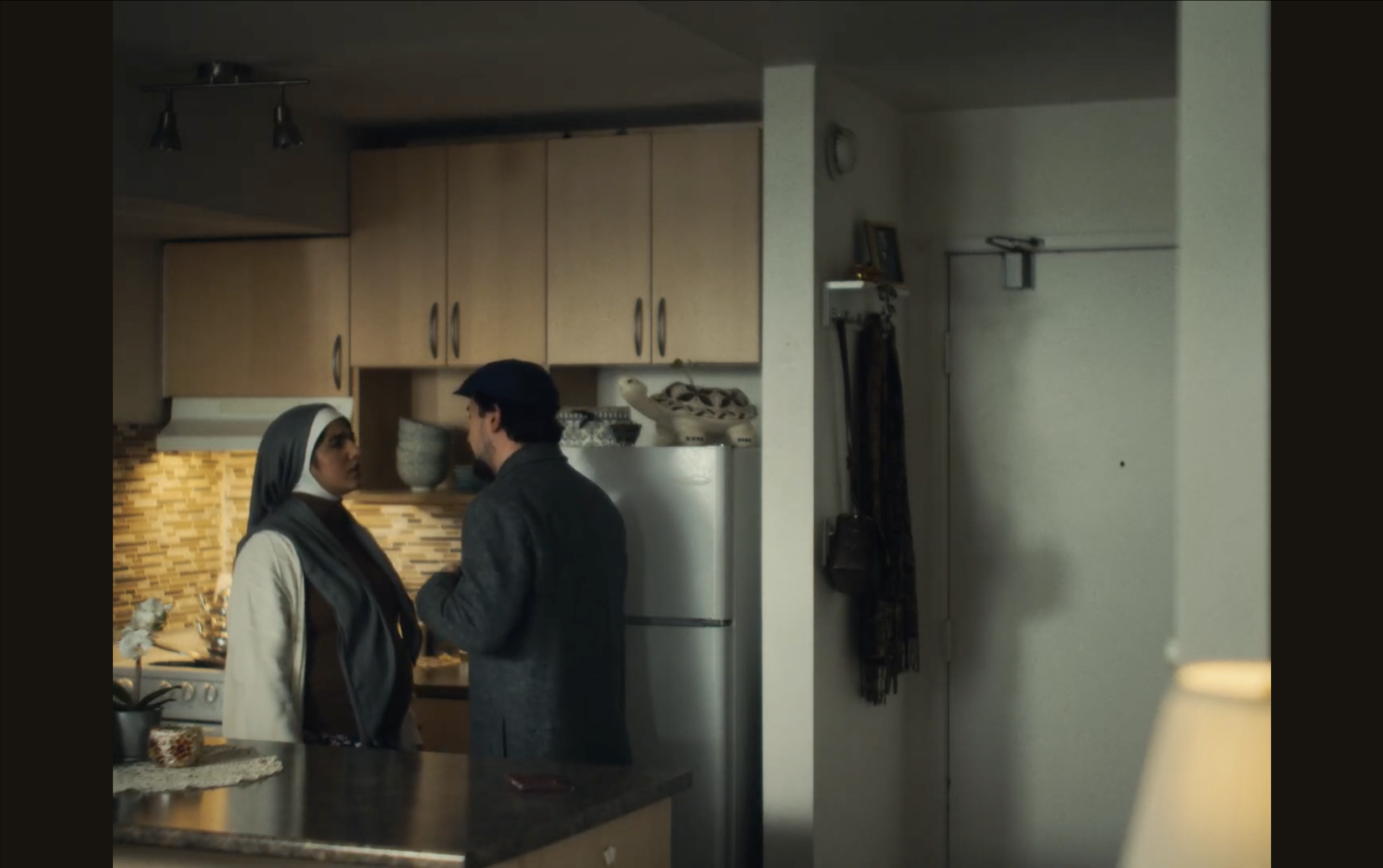 A woman and a man dressed in a dark jacket are standing close together in a kitchen, facing each other.