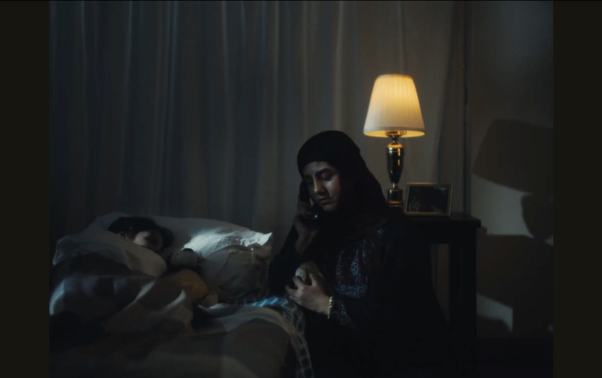 A woman sitting on a bed, looking down, in a dimly lit room with a bedside lamp, a framed photo, and curtains in the background.