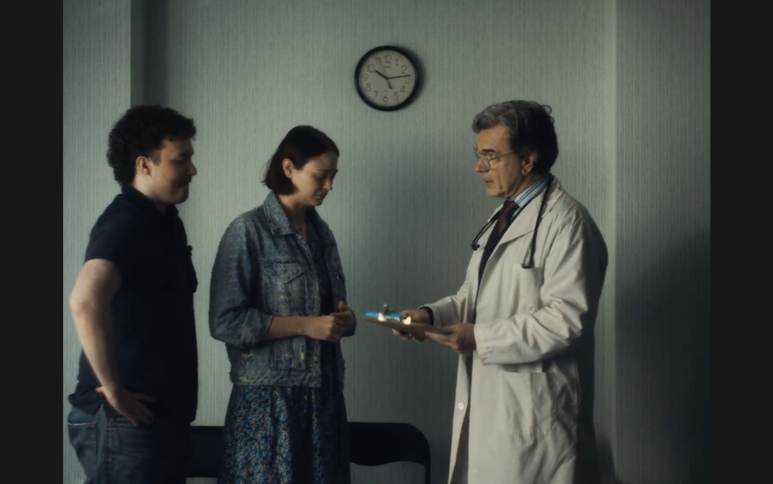 A male doctor in a white coat with a stethoscope around his neck presenting a tablet to two patients, a man and a woman, in a medical office. The wall behind them has a clock showing approximately 9:24.