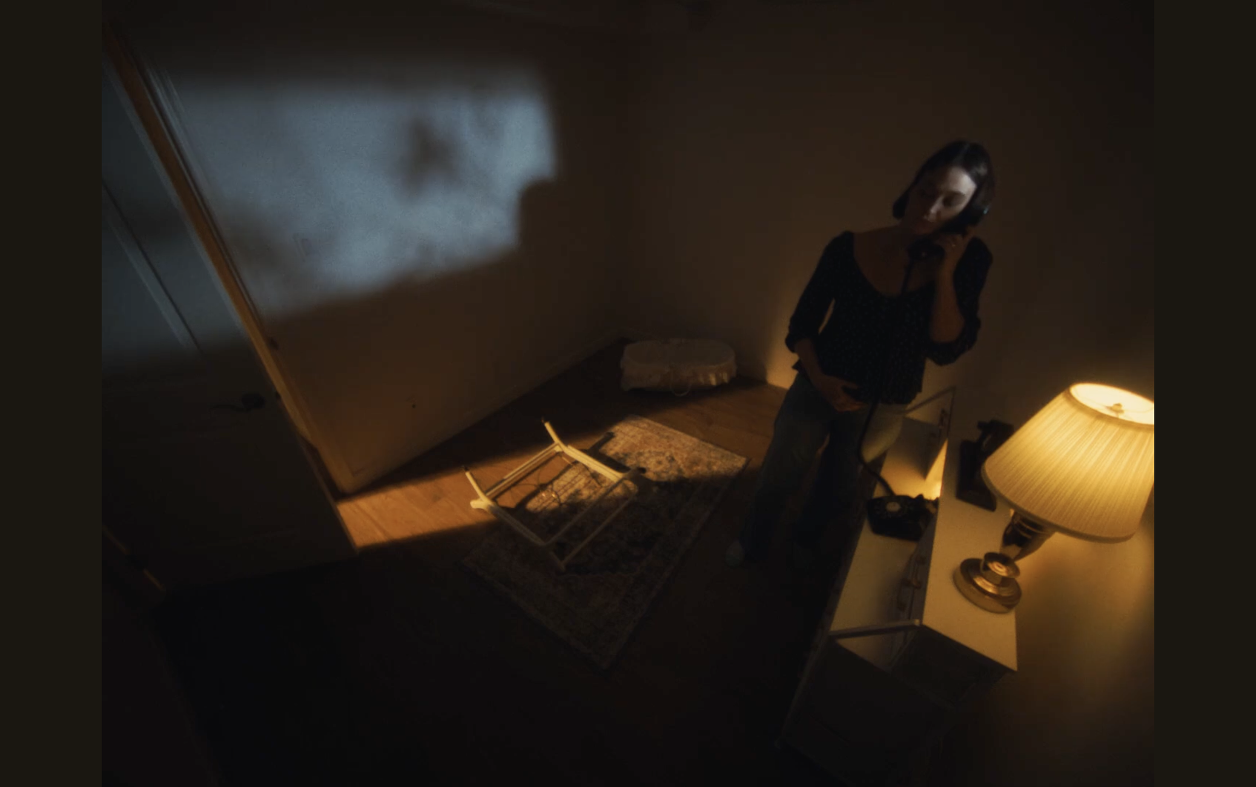 A woman with short dark hair stands in a dimly lit room. The room has a lamp on a side table, a rug, a few scattered objects, and a bassinet with a pillow in the background. The woman appears to be speaking into a phone.